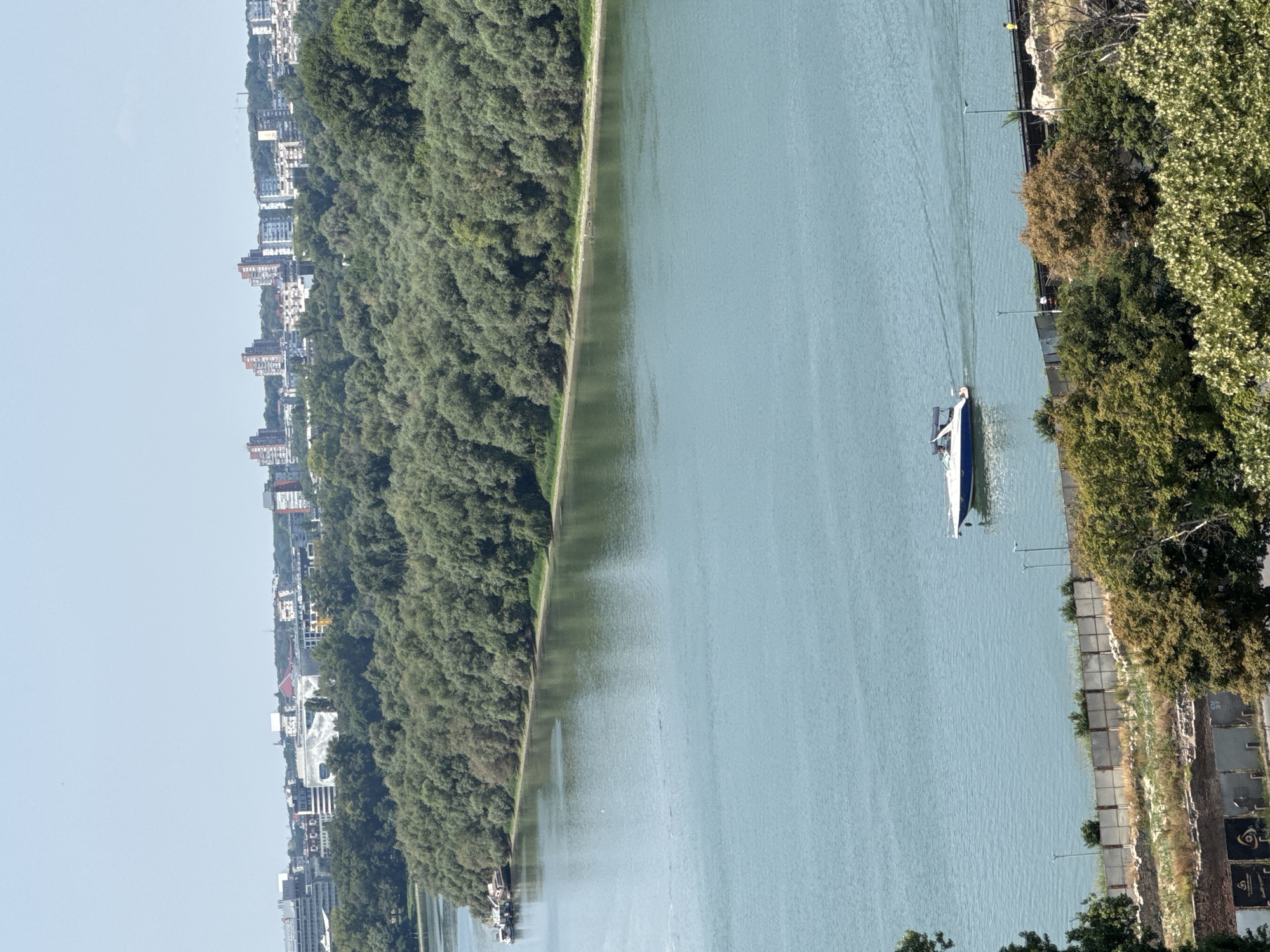 A wide, turquoise river flows through a city landscape, bordered by a dense forest on one side and urban buildings in the distance. A sleek, white and blue motorboat is navigating the river, leaving a faint wake behind it. The riverbank on the left is lined with a few docked barges. The background reveals a cityscape with various buildings of different heights and architectural styles, under a clear, bright sky. The foreground shows a more rugged, vegetated embankment with remnants of old structures, suggesting a historical or industrial area. The overall impression is a serene day with a blend of nature and urban development in Belgrade, Serbia.