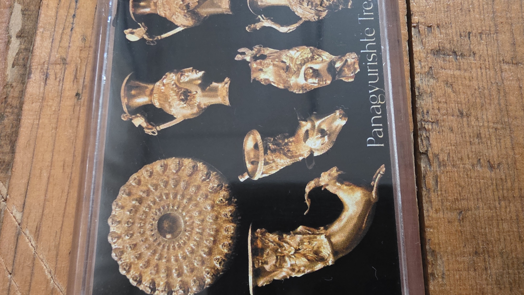 The image shows a photograph of a collection of golden artifacts, potentially of historical or archeological significance. The artifacts are displayed on a black background with the text "Panagyurishte Treasure" visible on the right side. The artifacts include what appear to be rhytons (drinking vessels) shaped like animal heads, and a flat, patterned golden object resembling a rosette or disc. The photograph is presented in a clear plastic frame and rests on a weathered wooden surface. The Panagyurishte Treasure is a Thracian gold treasure discovered in the town of Panagyurishte, Bulgaria, which is located in the Pazardzhik Province. Blagoevgrad is another city in Bulgaria, located southwest of Panagyurishte.