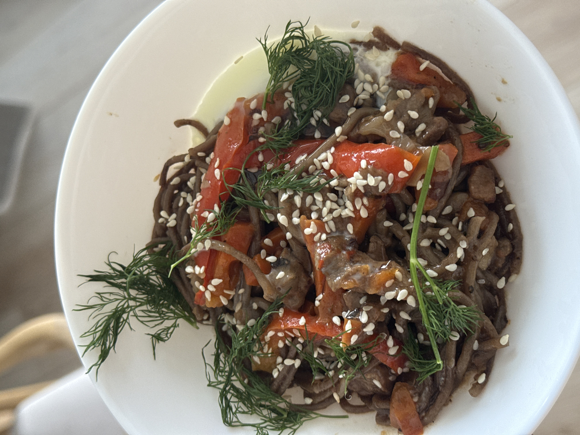 A top-down shot depicts a white bowl filled with a noodle dish. The noodles are dark brown, possibly soba noodles. Interspersed with the noodles are pieces of cooked red bell pepper and what appears to be either mushrooms or pieces of meat. The dish is garnished with sprigs of fresh dill and sesame seeds are scattered throughout. A light-colored sauce is visible in the background of the bowl. The edge of the bowl is visible on the left side of the image. The setting is indoors, with the edge of a chair visible. Location is unspecified and cannot be determined.