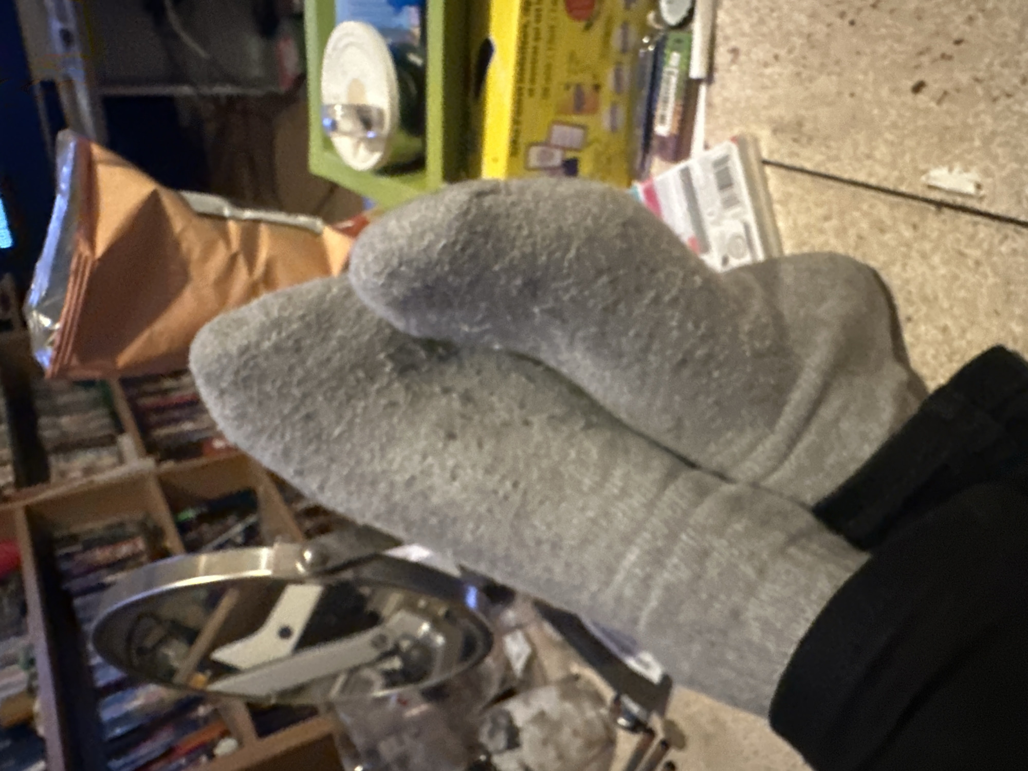 The image shows a pair of gray socks crossed over each other, with the cuffs visible, worn on a leg clad in black pants. In the background, there's a shelf filled with what appears to be DVDs. There is a brown bag of chips, and also a metal contraption. The floor is carpeted. There is no information to suggest that this image was taken in Gif-sur-Yvette, France.