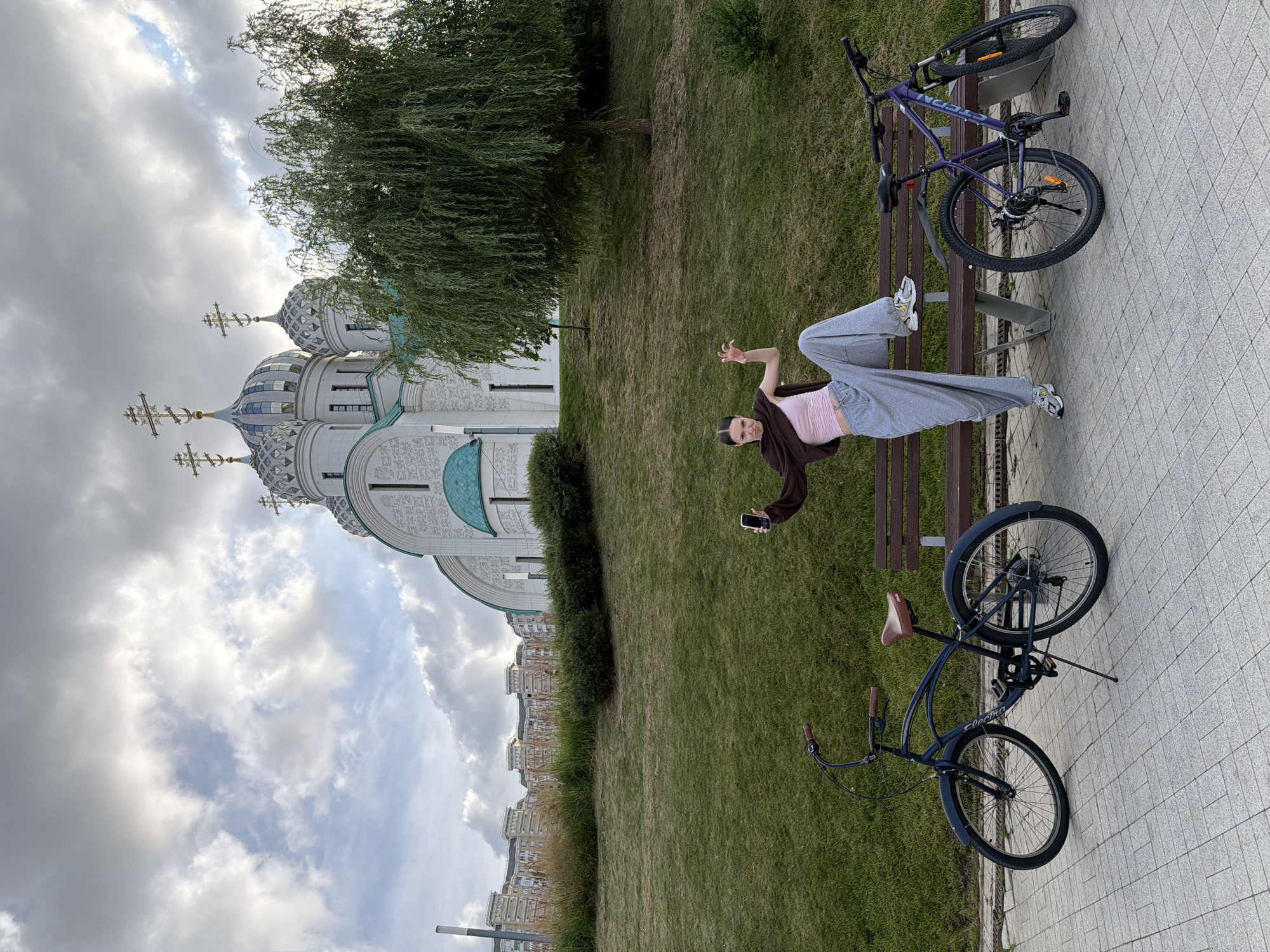 A young woman is sitting on a park bench next to a purple bicycle, holding her phone and appearing to take a selfie. She is wearing a light pink crop top, gray sweatpants, and white sneakers. Behind her, a large, domed building with crosses on top is visible, suggesting a religious or historical landmark. The building is white with green accents and intricate detailing. The scene is set in a park with lush green grass and a large weeping willow tree. Another bicycle, a dark blue cruiser, is parked on a tiled walkway. The sky is overcast with scattered clouds, indicating a possibly cool or pleasant day. The overall mood seems relaxed and casual, with the woman posing playfully. The location is Putilkovo, Russia. No visible text is present on the woman or the bicycles.