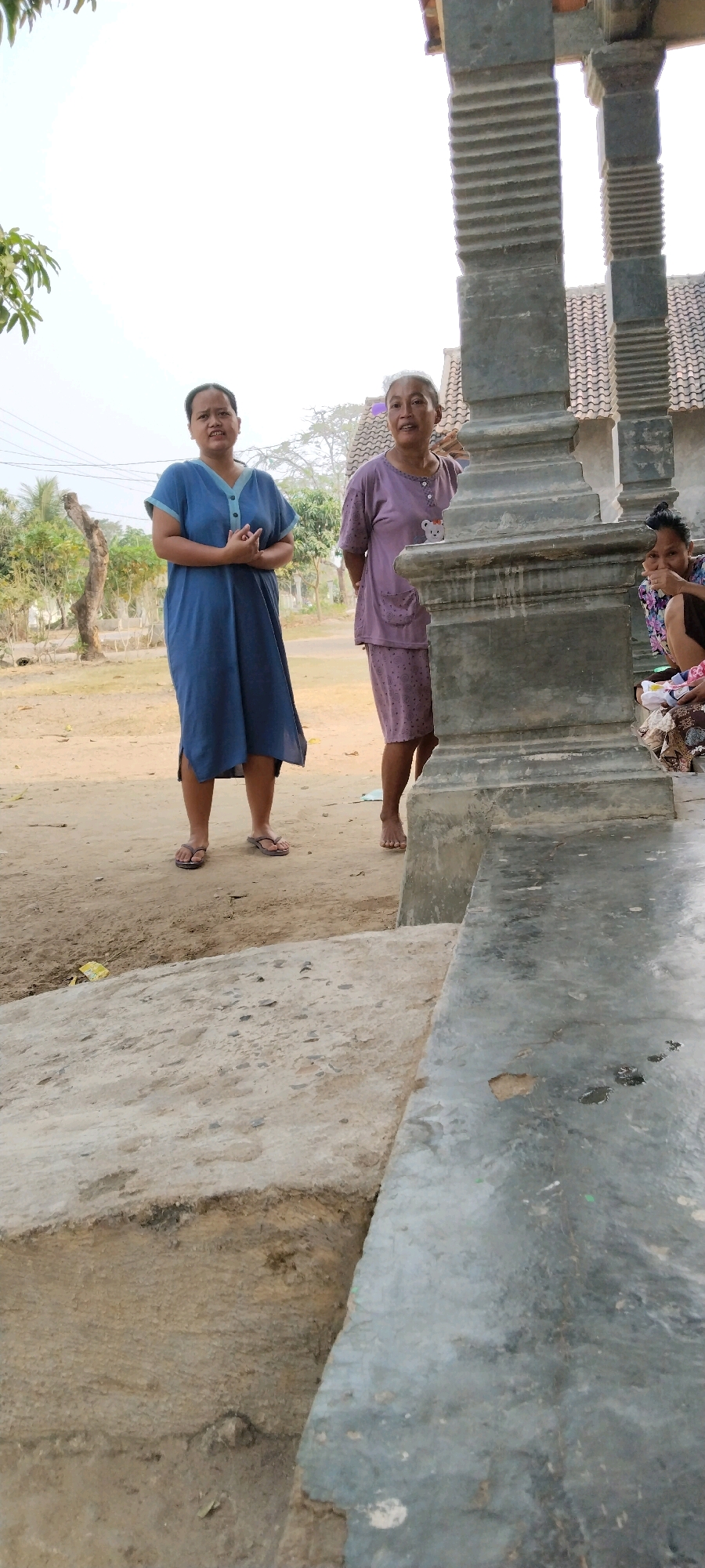 The image shows an outdoor scene in Tanjung Ratu Ilir, Indonesia. In the foreground, a textured concrete slab transitions to a dirt path. Behind this, two women stand near a large, weathered stone pillar. The woman on the left wears a blue dress, and the woman on the right wears a purple patterned outfit and a white head covering. A third person is partially visible to the right, crouching and looking towards the viewer. In the background, trees and a building with a tiled roof are visible. The overall lighting suggests it is daytime.