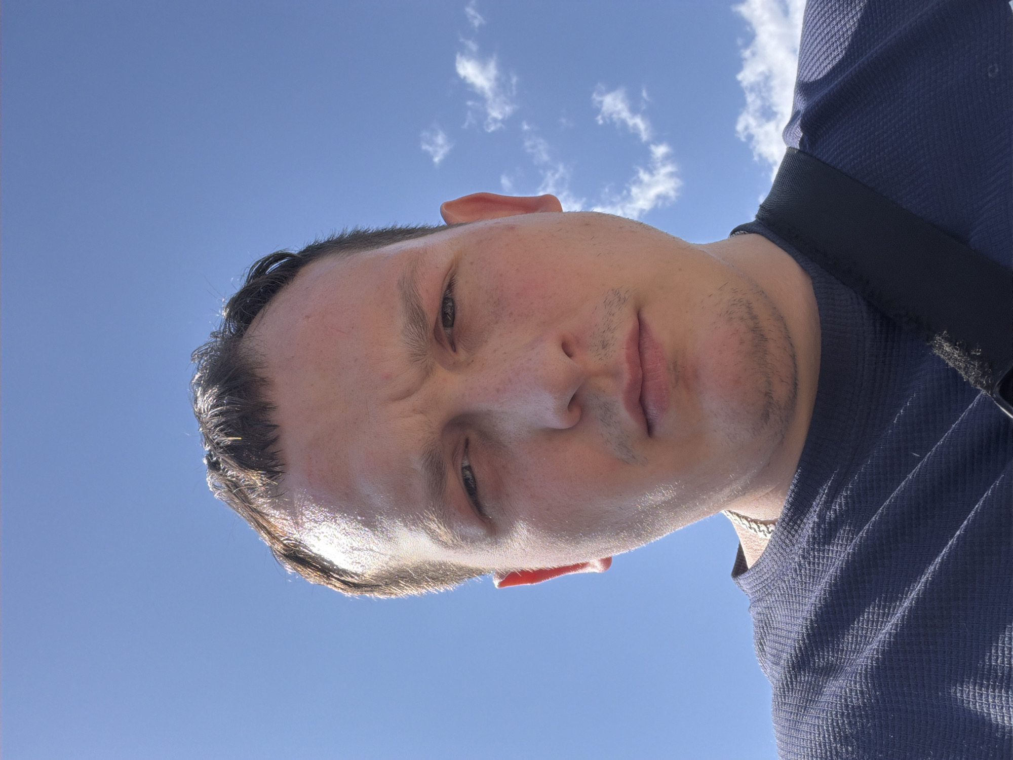 A close-up, low-angle portrait of a man against a bright blue sky with scattered white clouds. The man is light-skinned with short, dark hair, a stubbly beard, and a slightly furrowed brow. He wears a dark blue waffle-patterned shirt with a black strap running across his right shoulder. A silver chain necklace is visible at his neck. The picture was taken outside on a sunny day. I do not have the information about where the photo was taken in Chaikovsky, Russia.
