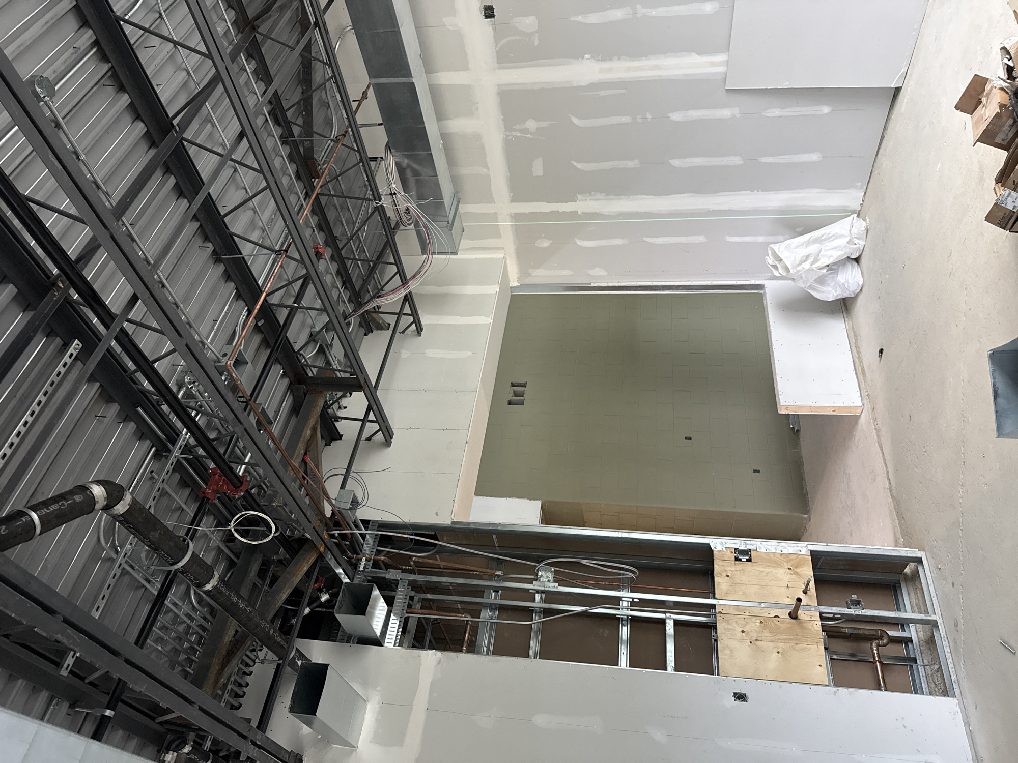 The image depicts the interior of a building under construction in Brampton, Canada. The scene is characterized by exposed building materials, including metal framework, ductwork, plumbing, and electrical wiring suspended from the ceiling. Drywall is installed on the walls, with visible seams and joint compound, indicating that finishing work is in progress. The floor is a bare concrete slab. In the foreground, a section of exposed framing reveals pipes and conduits. To the right, two large white bags, possibly containing construction debris or materials, are present. In the background, an open doorway reveals a room with cinder block walls. The overall impression is one of an unfinished commercial or industrial space.