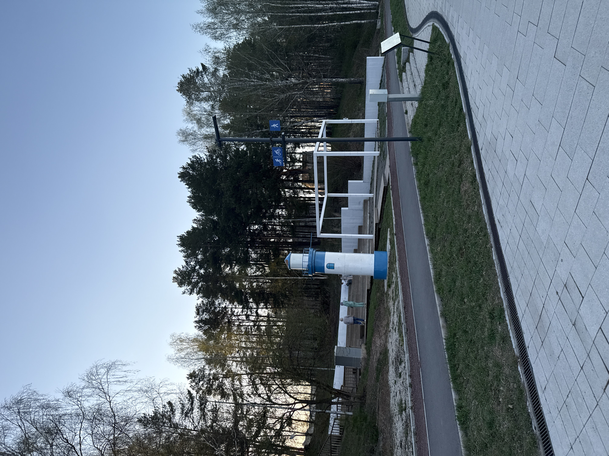 The image shows a park-like setting with a model lighthouse. The sky is pale blue. A dense forest forms a backdrop. The lighthouse is painted white with blue accents on top and bottom. A walking/biking path runs alongside the lighthouse. A signpost with blue pedestrian and bicycle symbols is visible. A paved area is present on the right side of the image. Two people are standing by the lighthouse. The location is in an unknown city and unknown country.