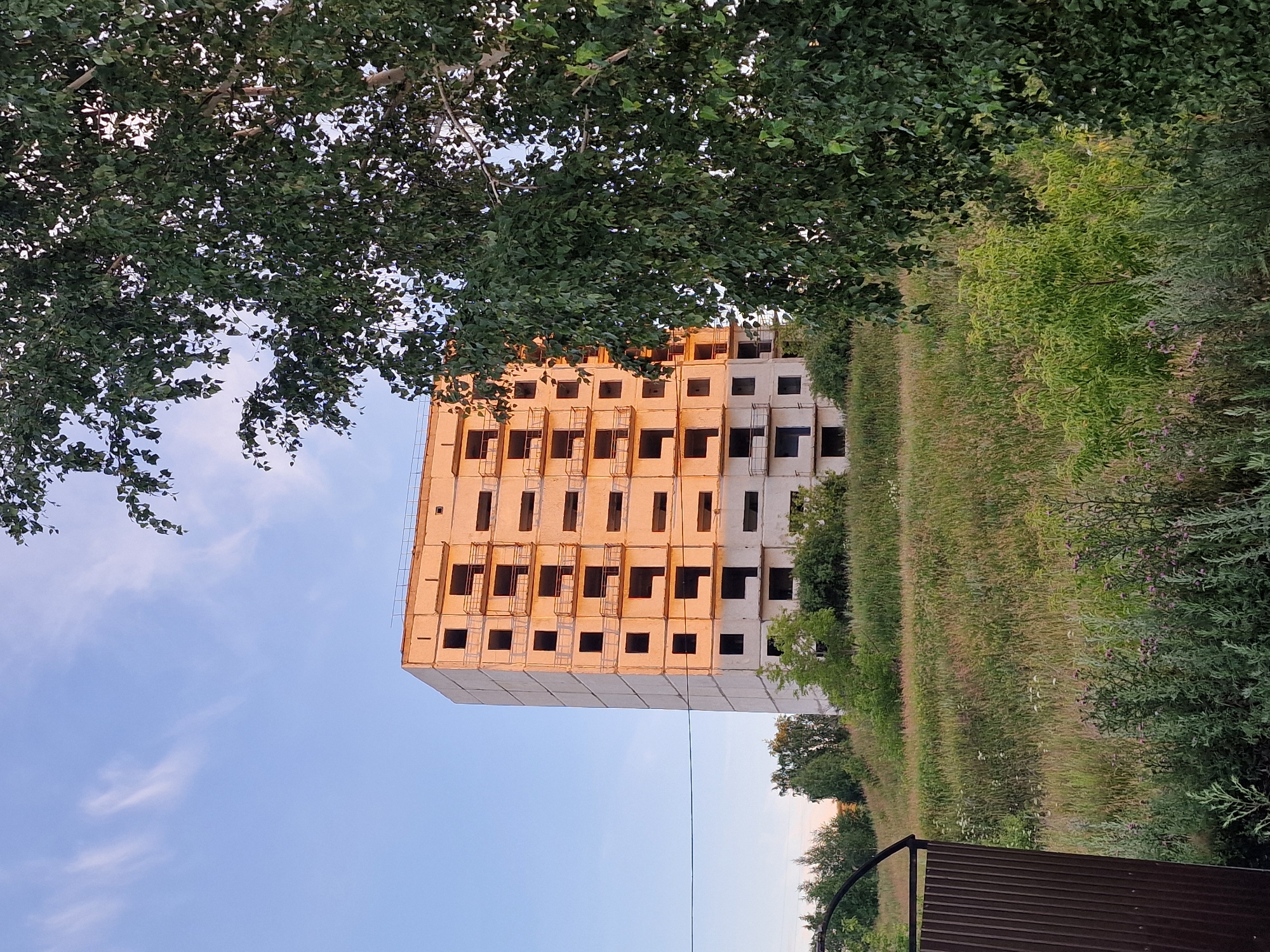 The image depicts an unfinished, multi-story concrete building under a clear sky with sparse, wispy clouds. The building, which appears to be residential, has numerous dark, empty window openings and exposed balconies. It is situated on a grassy hillside with scattered trees and vegetation. A tall, leafy tree is prominent in the foreground on the right side of the frame, partially obscuring the building. A metal gate, made of corrugated material, is visible on the left. The lighting suggests it might be late afternoon or early morning, with the side of the building illuminated by warm sunlight. There are no people visible in the image. The scene appears to be in a semi-rural or developing urban area. The location is identified as Kashgar City, China.