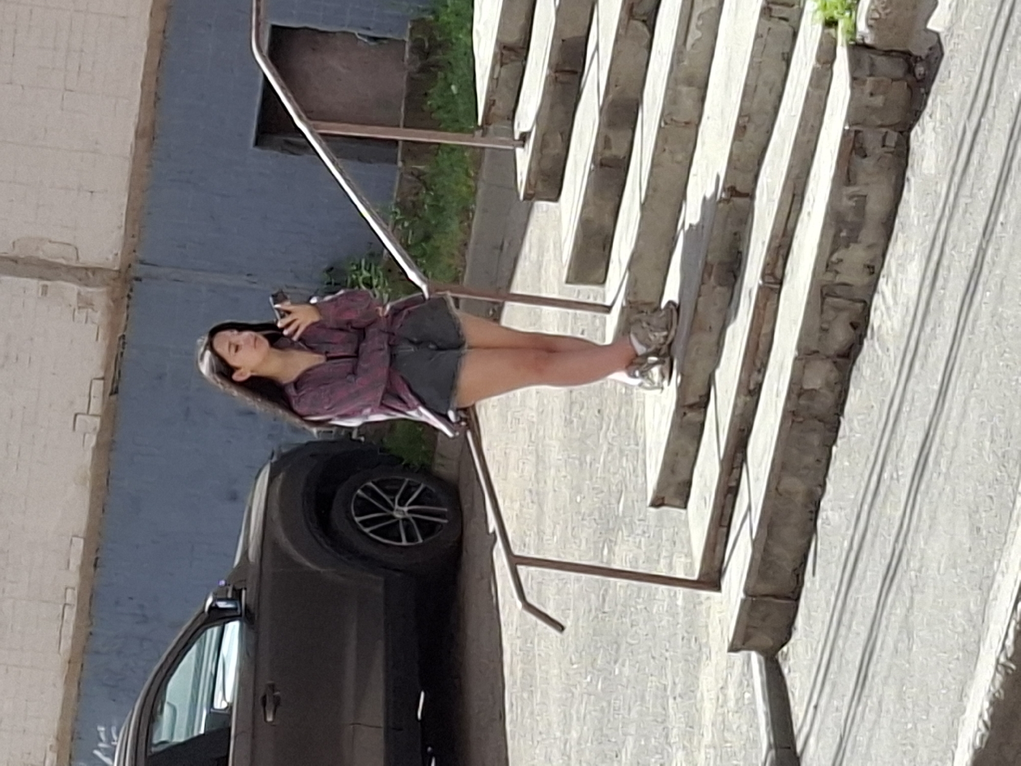 A young woman stands on concrete stairs, holding a black mobile phone in her right hand. She is wearing a pink and purple plaid shirt, a black tank top, and grey denim shorts. Her feet are clad in tan and white sneakers. The stairs have metal railings on either side. Behind her, a dark-colored SUV is parked. The environment appears to be an urban setting with a blue and white building in the background. The lighting suggests it is daytime.