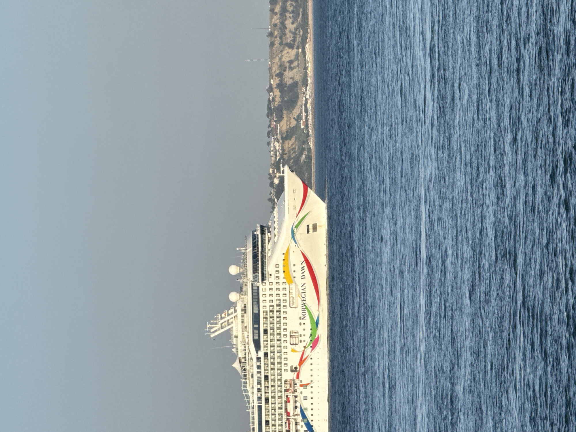 A large white cruise ship, the "Norwegian Dawn," sails on a calm blue sea. The ship features a colorful, wavy design along its hull and has multiple decks with visible windows. In the background, a coastal town with houses and some vegetation is situated on a hillside. The sky is a clear, pale blue. The scene appears to be a sunny day. The location is the coast of Oeiras, Portugal, as indicated by the distinctive coastline visible behind the ship.