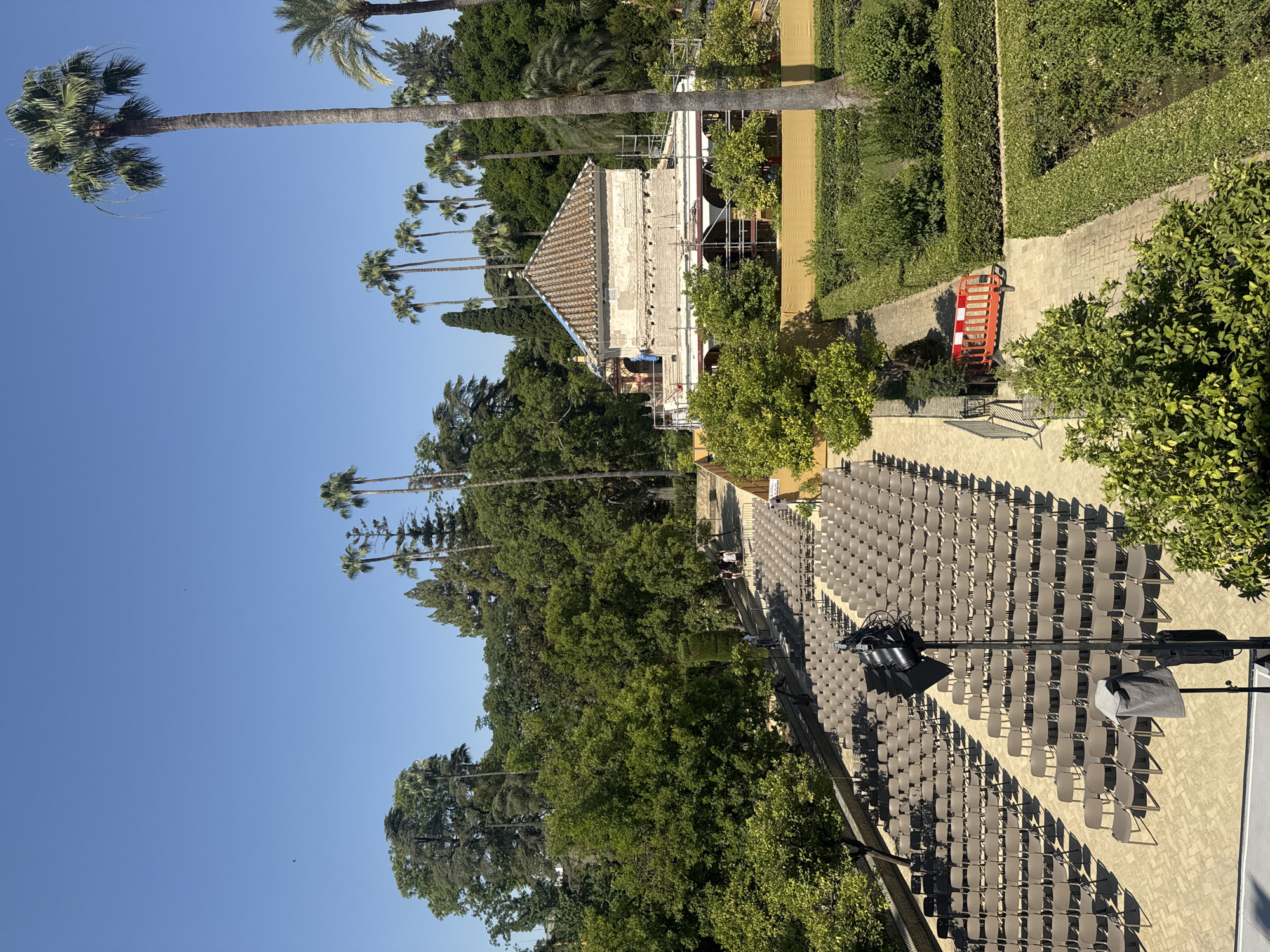 This is an outdoor scene in the gardens of the Alcázar of Seville, Spain. In the foreground, a large area is set up with rows of empty brown chairs, suggesting a performance or event space. A tall speaker on a stand is positioned in front of the chairs. Beyond the seating area, a pathway leads towards a building that appears to be undergoing renovations, with scaffolding around it. The building has a tiled roof and a pale facade. The background is filled with lush greenery, including numerous palm trees and other trees, under a clear blue sky. Several people are visible walking in the distance, and some are near the building. A orange construction barrier is placed on a path.