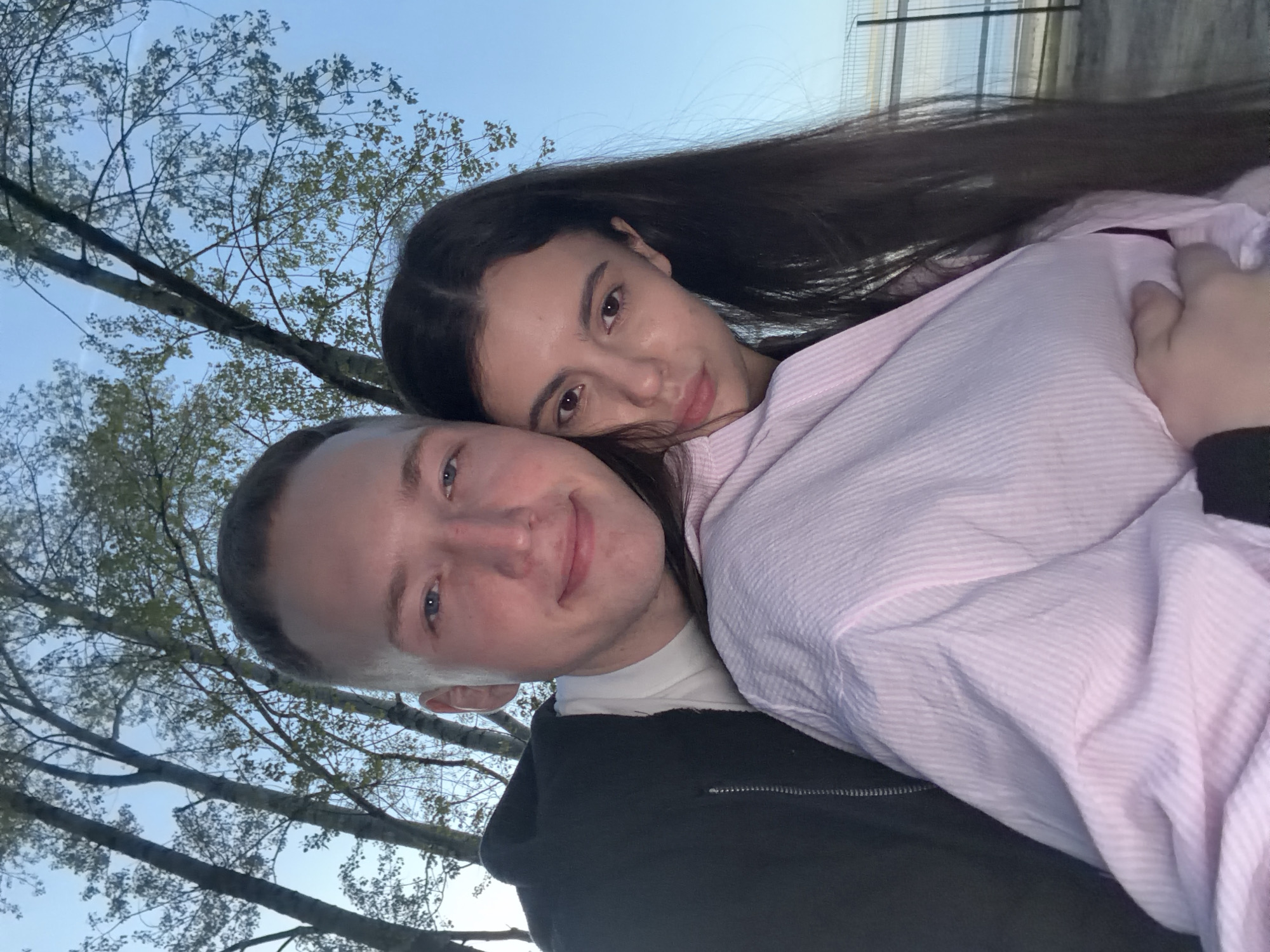 A selfie-style, outdoor photograph captures a young couple embraced. The man is wearing a black jacket over a white undershirt. The woman has long, dark hair and a light pink striped shirt. Branches of bare trees, typical of a temperate climate, are visible in the background against a light blue sky. A building, possibly a greenhouse or similarly structured edifice, is partially visible in the upper right corner of the frame. The location might be a park or public garden area in Chaikovsky, Russia, given the presence of natural elements and building structure.