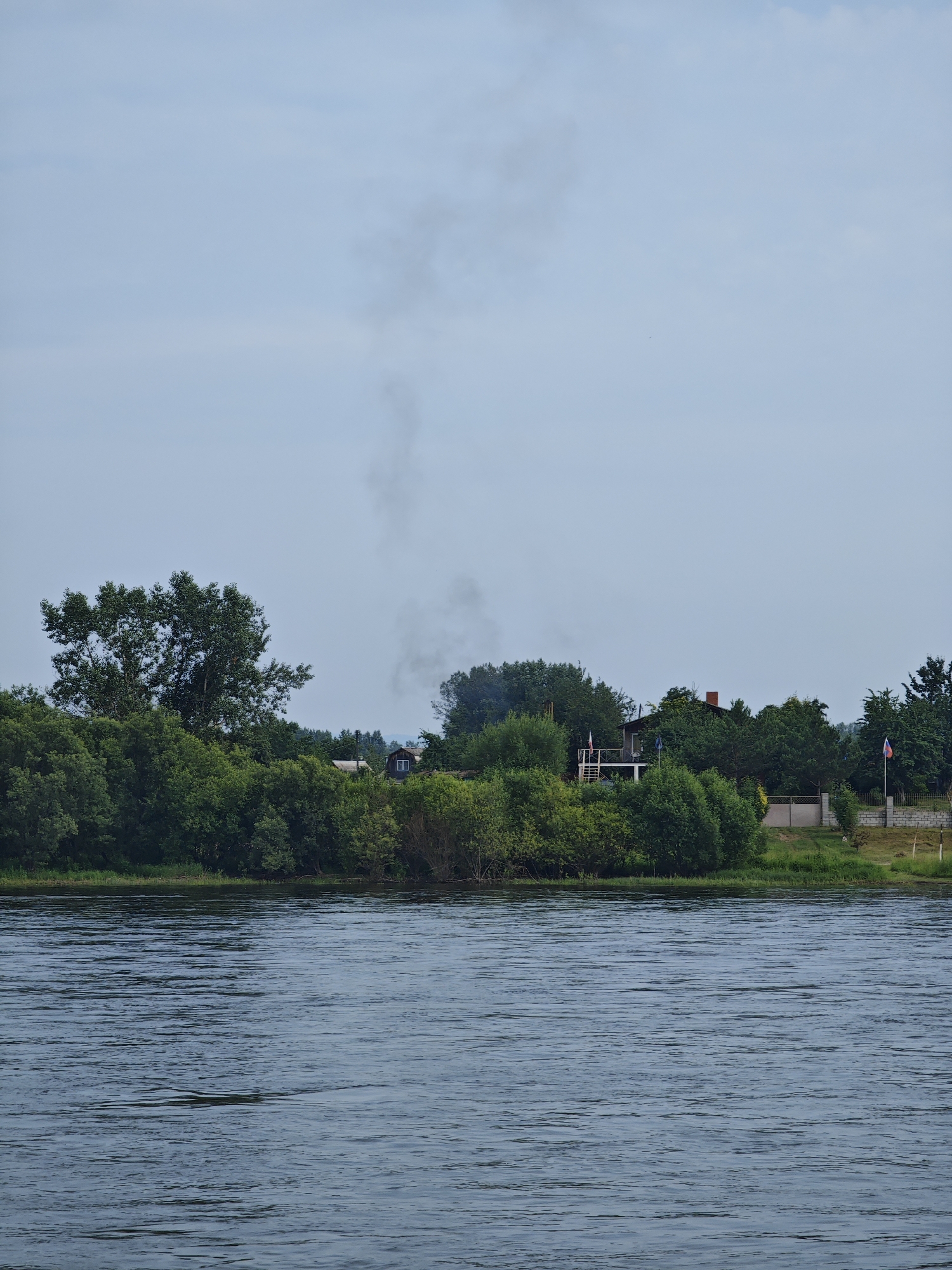 The image depicts a wide river in the foreground, with gentle ripples on its surface. The riverbank is lined with lush green trees and dense foliage. In the background, several houses are visible amongst the trees, suggesting a rural or semi-rural settlement. A plume of dark smoke rises from behind the trees on the left side of the image, indicating a fire or chimney activity. A flag on a flagpole is also visible on the right side of the image, near a fence and a building. The sky is overcast with light clouds, and the overall atmosphere appears calm and serene. The location is identified as being in the city of Sosnovoborsk, Russia, which aligns with the natural landscape and architectural style suggested by the image.