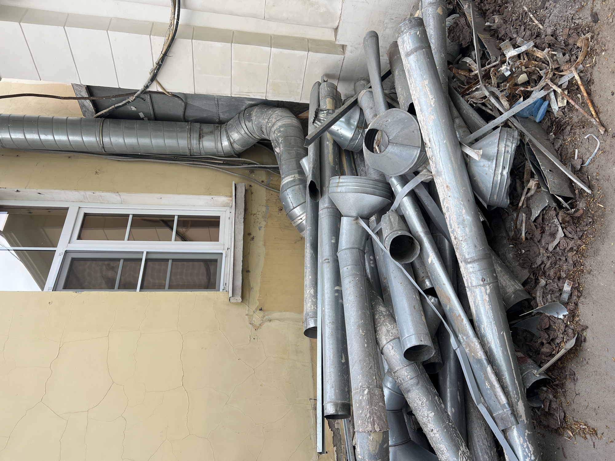 The image shows a pile of galvanized metal pipes and fittings stacked against a yellow building with a white-framed window. The pipes appear to be used and possibly discarded, mixed with leaves and some metal scraps. A section of similar metal ductwork is affixed to the building, running horizontally. The setting appears to be an exterior space, possibly an alley or the side of a building. There is no explicit information indicating that the location is Moscow, Russia.