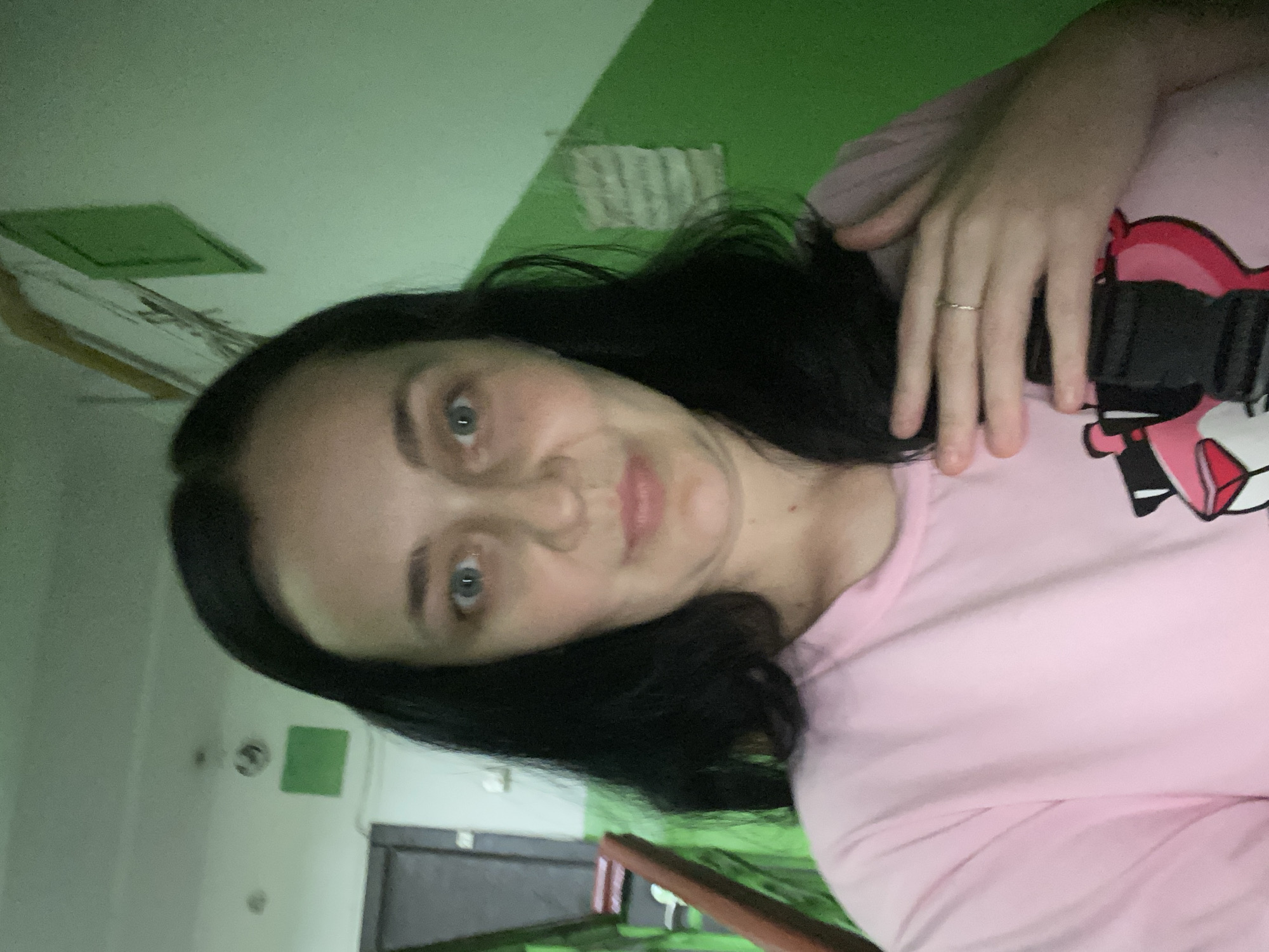 A woman with dark hair is taking a selfie in a dimly lit hallway. She is wearing a pink t-shirt with a cartoon character on it. Her right hand is resting on her chest. The hallway has green walls and a wooden railing on the left. There are also some posters or notices on the walls.