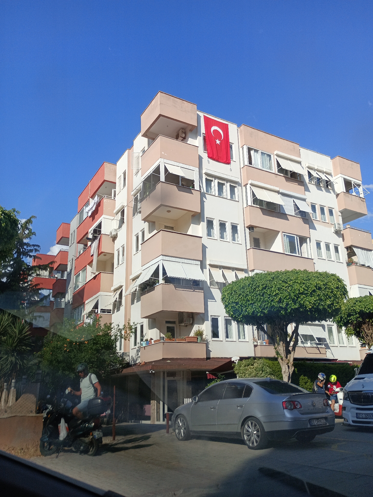 A wide shot captures a modern apartment building in Alanya, Turkey. The building has multiple balconies and is painted in shades of cream, beige, and coral. A large Turkish flag with a white crescent moon and star hangs from one of the balconies, illuminated by bright sunlight. Below the building, a man on a scooter is parked on the side of the road, wearing a light blue t-shirt and shorts, with a backpack on the back of his scooter. A silver sedan is parked in front of the building, and another car, possibly a white van, is parked to the right, partially visible. Two people, one wearing a yellow helmet and the other a blue helmet, are standing next to a scooter in front of the white vehicle. A large, round, leafy tree stands prominently in front of the apartment building, adding a touch of greenery to the urban scene. The sky is a clear, vibrant blue, suggesting a sunny day. The overall impression is one of a typical day in a Mediterranean coastal city, with residential buildings, vehicles, and people going about their routines. The Turkish flag is a clear indicator of the national identity of the location. The image appears to have been taken from a vehicle, as indicated by the reflection on the bottom of the frame.