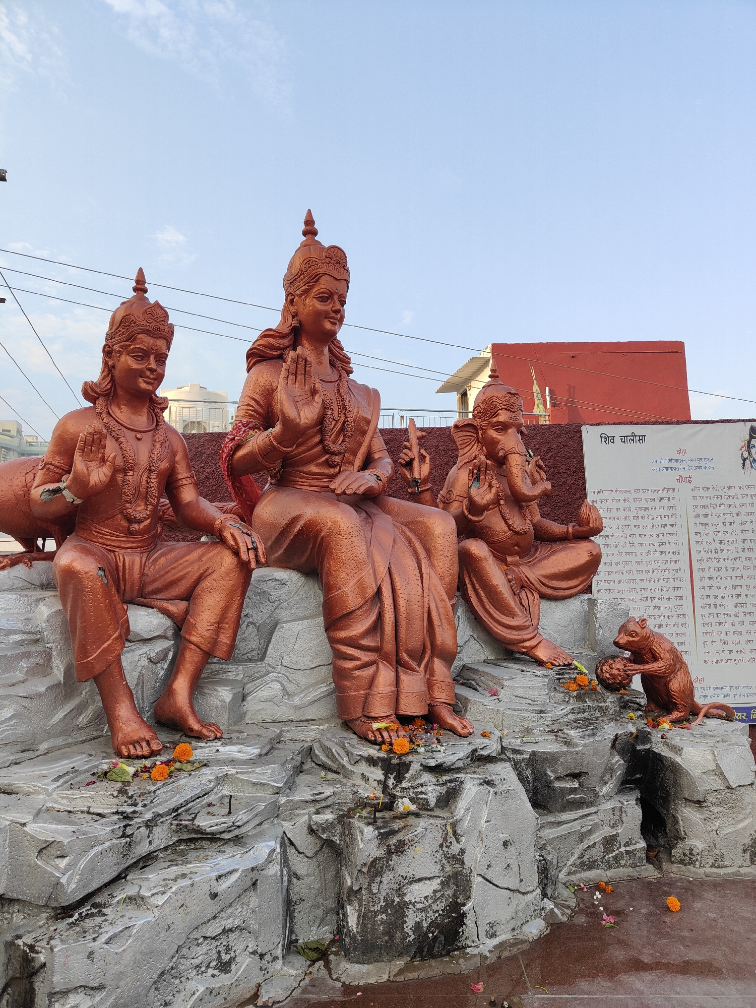 This image depicts a statue of Hindu deities, likely in Gurgaon, India. The central figure, a goddess with multiple arms, is seated. To her left, a male deity is seated, and to her right, Ganesha, the elephant-headed god, is also seated. In front of Ganesha, a small statue of a monkey is shown holding a round object. The statues are made of a bronzed material and are placed on a rocky, textured base. Several orange marigold flowers are scattered around the base of the statues, suggesting a religious offering. In the background, a red building and some power lines are visible, along with a clear blue sky. A large white banner with text in Hindi is partially visible behind Ganesha and the monkey statue. The overall scene suggests a public shrine or a religious monument.