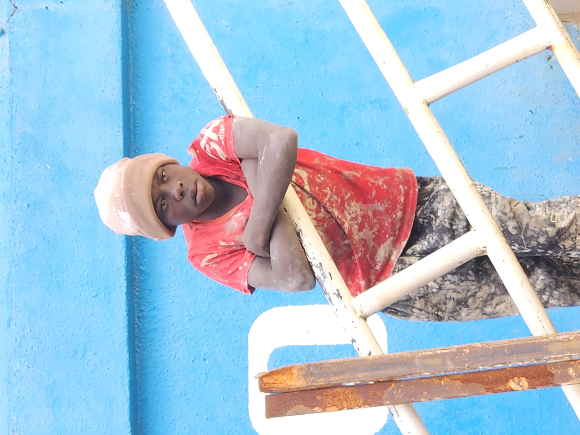 A young Black male is standing and leaning on a white railing against a bright blue textured wall. He is wearing a light brown beanie, a red t-shirt splattered with white paint, and camouflage pants. His arms are crossed in front of him, and his hands are resting on his biceps. He is looking directly at the camera with a serious expression. To the left of the male, two rusted metal bars are leaning against the blue wall. The railing appears to be part of a set of stairs or a walkway. The overall scene suggests an outdoor urban environment, possibly a construction or industrial area, given the paint splatters and rusted metal.