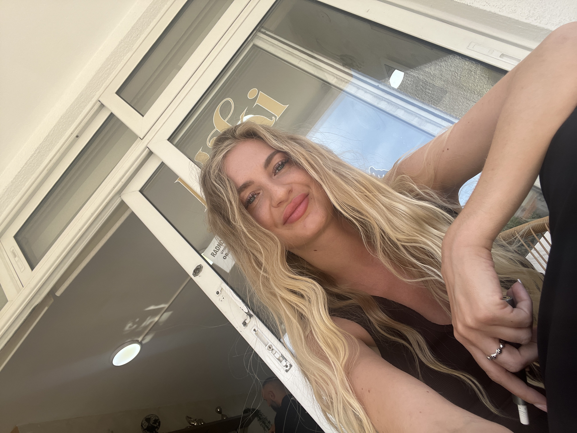 A woman with long blonde wavy hair is posing for a selfie in front of a large glass window/door, likely in Budva, Montenegro. The window has a white frame and gold lettering visible on the glass. The woman is wearing a dark brown top and a ring on her finger. Inside, behind the window, a man's head is visible in the lower frame. The background shows a ceiling with a light fixture and part of a decorative object is visible in the lower corner.