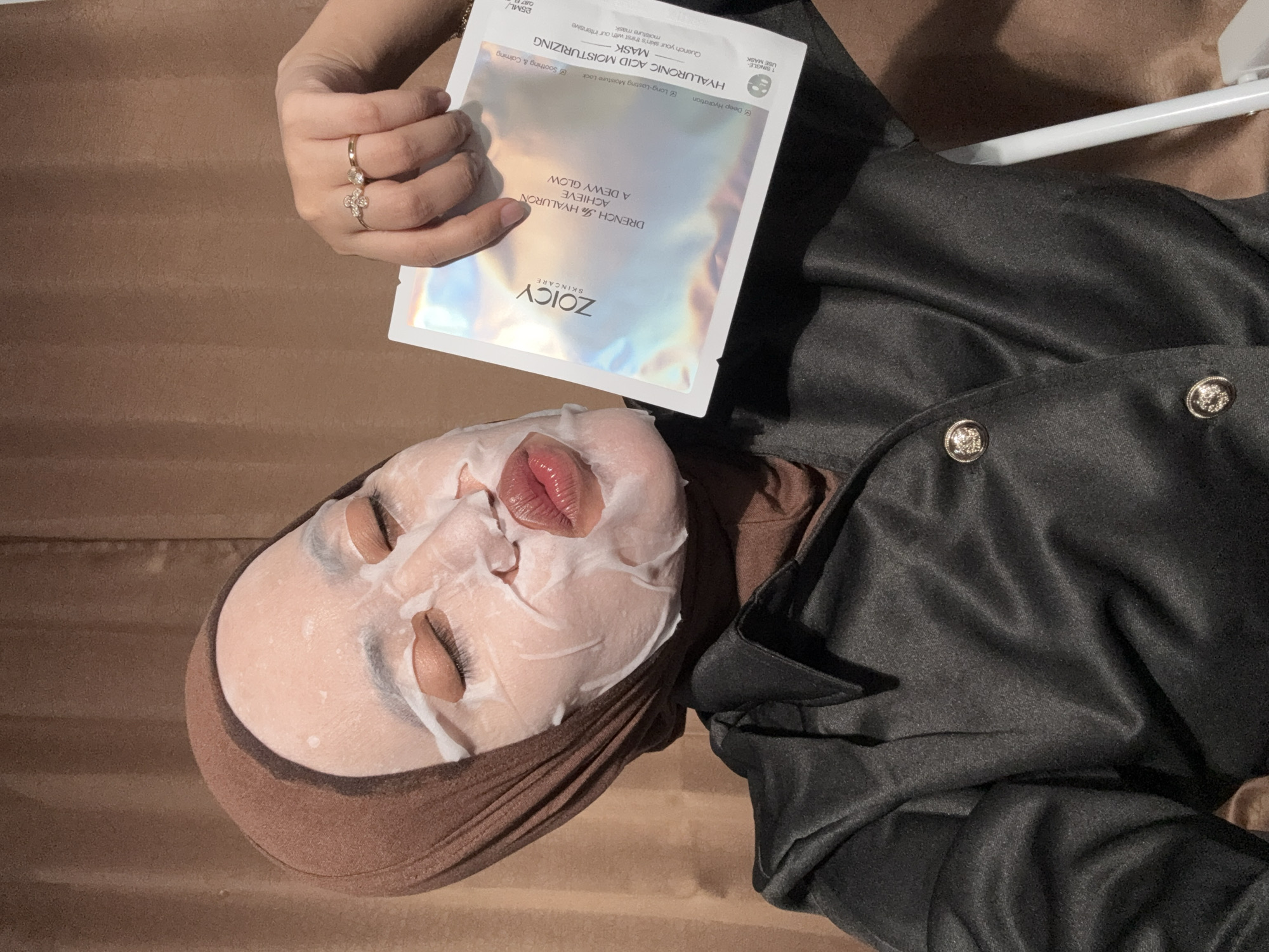 An overhead shot captures a person relaxing indoors in Gadingrejo, Indonesia, while applying a skincare sheet mask. The individual is lying down, with a white, translucent sheet mask covering most of their face, leaving the eyes and lips visible. Their eyes are closed, indicating a state of rest or relaxation. A brown head covering or hijab frames their face.
A hand in the upper left holds a silver, iridescent skincare product package. This package is clearly branded "ZOICY SKINCARE" and identifies the product as a "HYALURONIC ACID MOISTURIZING MASK." The packaging prominently states the product's benefits: "DRENCH In HYALURON, ACHIEVE A DEWY GLOW," offering "Deep Hydration," "Long-Lasting Moisture Lock," and "Soothing & Calming." It is a "1 SINGLE USE MASK" containing "25ML / 0.87 fl. oz," designed to "Quench your skin's thirst with our intensive moisture mask." The hand holding the package is adorned with two elegant rings, one featuring a decorative floral or bow-like design with small gems, and another simple band.
The person is wearing dark-colored clothing, possibly a jacket or top, visible on the right side of the frame, which features several silver-toned decorative buttons. The background consists of a uniform brown surface, likely a bed or sofa, contributing to the tranquil setting. A white horizontal bar, possibly part of the furniture, is also visible on the right.