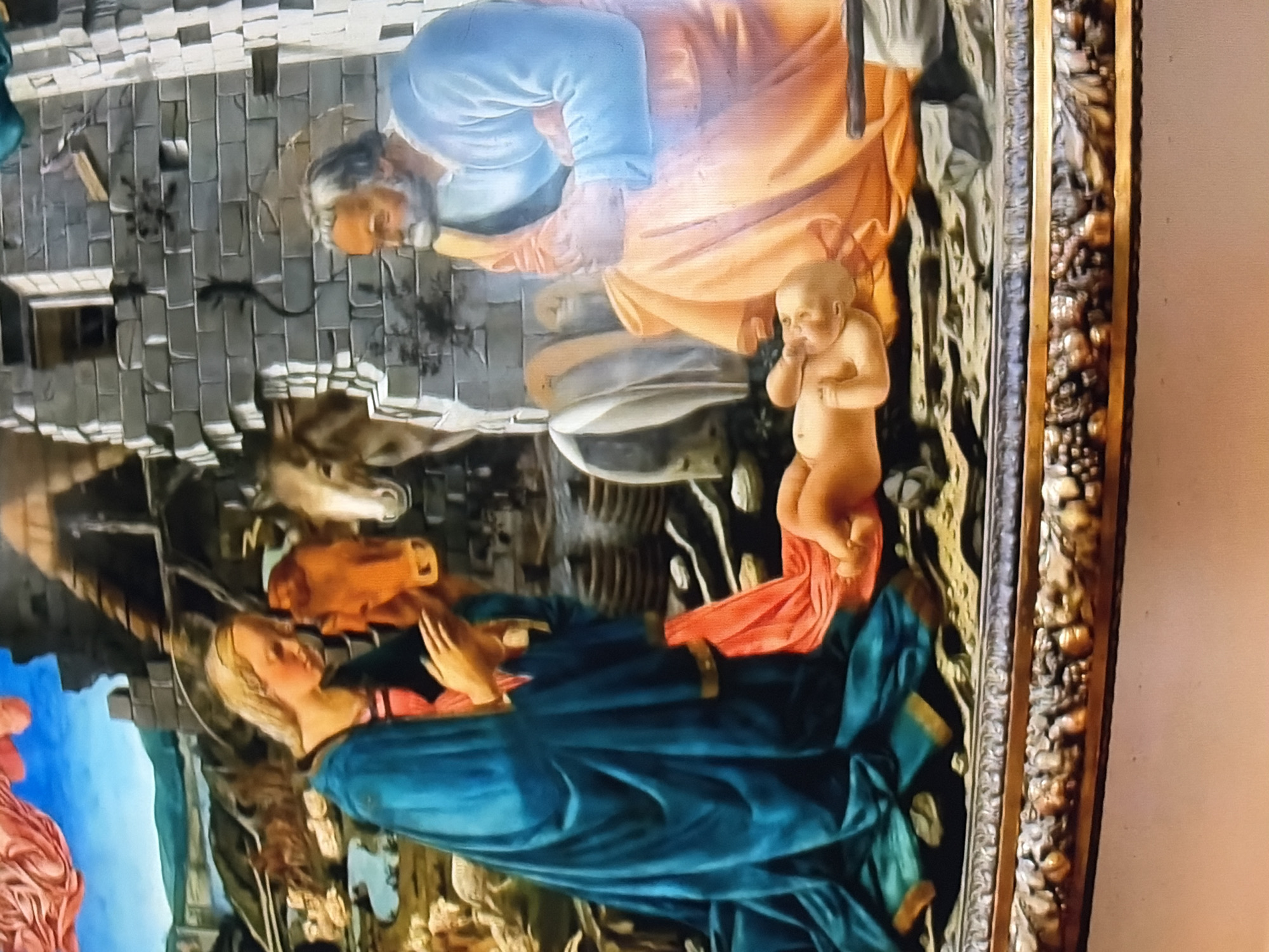 The painting depicts a Nativity scene with figures of Mary, Joseph, and the baby Jesus. Mary, dressed in a blue mantle and red undergarment, is kneeling with her hands clasped in prayer. Joseph, wearing an orange robe and blue garment, sits beside her, gazing at the infant. The baby Jesus is lying on straw, reaching towards his mouth. In the background, a stable structure is visible, with an ox and a donkey. The scene is framed by an ornate golden border. The location is described as Unknown, Unknown.