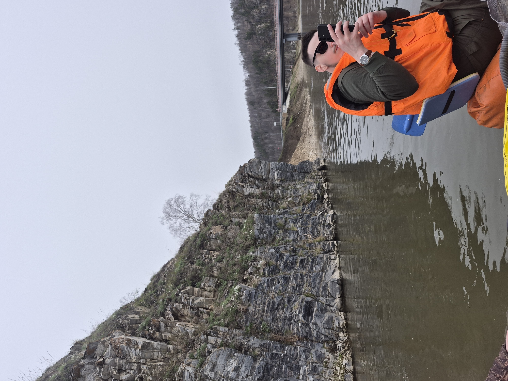 A man in a bright orange life vest, dark green long-sleeved shirt, and sunglasses is captured from the chest up, seemingly sitting in a boat or raft on a body of water. He holds a black smartphone with both hands, aiming it towards a rocky cliff face, appearing to take a photograph or video. A watch is visible on his left wrist. Adjacent to him in the boat is a blue floating pad, and a yellow inflatable edge is partly visible.
The scene features a prominent, steep rocky cliff on the left, rising directly from the water's edge. The cliff face displays distinct horizontal geological layers, with scattered patches of green vegetation and a single bare tree perched near its top. The water is calm, reflecting the pale, overcast sky and elements of the shoreline.
In the background, a long bridge with visible infrastructure spans across the water, connecting distant land areas. Beyond the bridge, rolling hills covered with what appear to be bare trees under an overcast sky complete the natural setting. The overall atmosphere suggests a cool, possibly early spring or late autumn day, likely taking place in an outdoor recreational setting near Новомунасипово, Russia. No visible text is present in the image.
