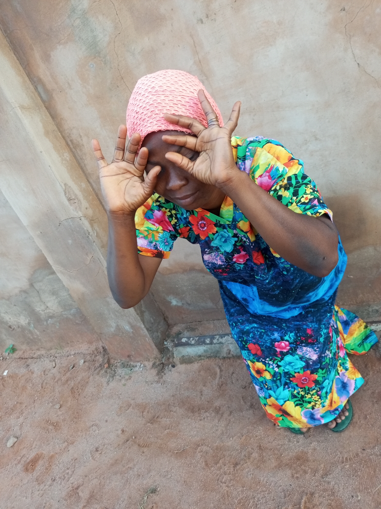 A medium shot from a high angle shows a woman wearing a pink headscarf and a brightly colored floral dress. She is kneeling on reddish-brown dirt in front of a textured, light brown wall. Her hands are raised to her face, covering her eyes, with her fingers spread apart in a playful or secretive gesture. The wall behind her has a rough, uneven surface and appears to be weathered. The ground in front of her is dusty and shows some footprints. The overall impression is of an outdoor scene, possibly in a rural or less developed urban area. The location is identified as Tempure, Nigeria.