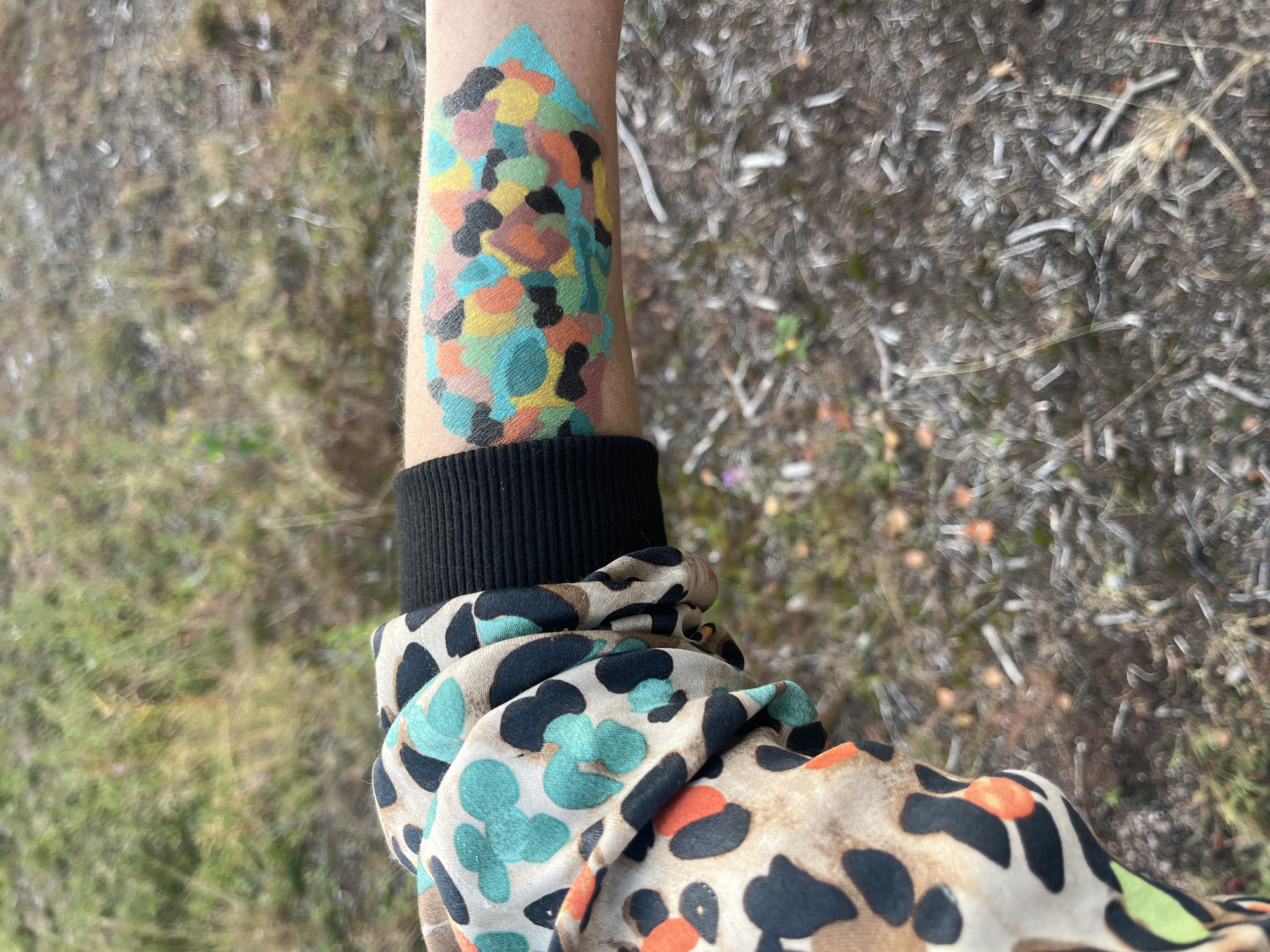 The image shows a close-up view of a person's forearm with a colorful, abstract pattern painted on it. The pattern consists of various irregularly shaped patches of teal, orange, yellow, pink, and black. The person is wearing a black cuff on their arm, and a portion of a garment with a leopard-like print in brown, black, and teal is visible on the left side of the frame. The background is out of focus and appears to be an outdoor environment with dry vegetation and soil. The lighting suggests it is daytime. No city or specific location is identifiable.