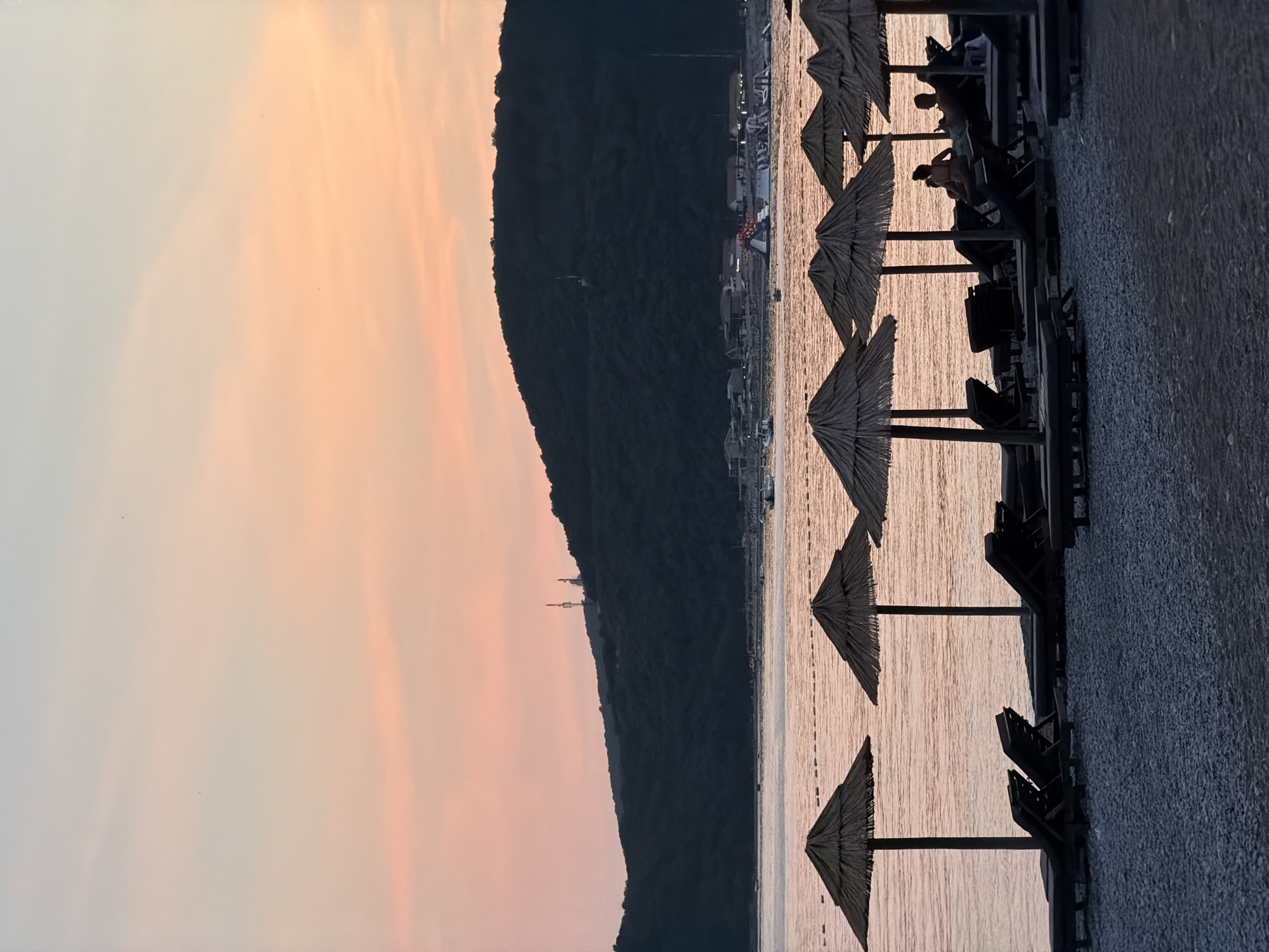 The image depicts a tranquil sunset scene on a pebble beach in Prijevor, Montenegro. In the foreground, several thatched umbrellas stand tall over empty lounge chairs, casting long shadows on the stones. A few figures are seated on lounge chairs, silhouetted against the soft pink and orange hues of the sky and the calm, reflective water. The water is dotted with a floating inflatable water park, and in the distance, a lush green hillside dominates the horizon, with faint structures and communication towers visible on its crest. Boats are scattered across the water, suggesting a leisurely end to the day. The overall atmosphere is one of peaceful serenity as dusk settles over the Adriatic coast.