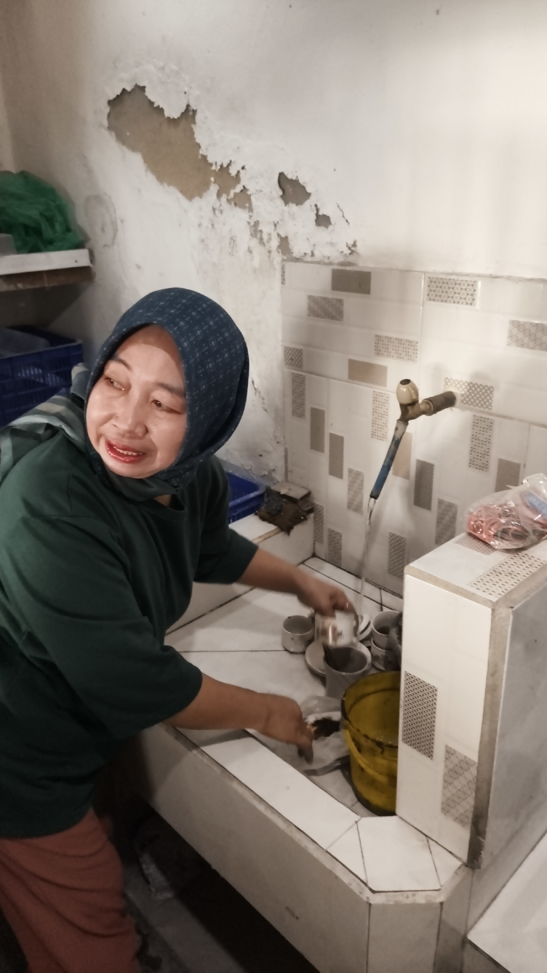 A middle-aged Indonesian woman wearing a blue patterned hijab and a green t-shirt is washing dishes in a sink. She is standing in a kitchen area with white tiled walls and a concrete countertop. The sink is filled with water, and she is holding a cup. There are other dishes in the sink, as well as a yellow bucket. The wall behind the sink has peeling paint. A faucet is visible, with water running from it. The woman appears to be smiling. The scene is set in Dalegan, Indonesia.