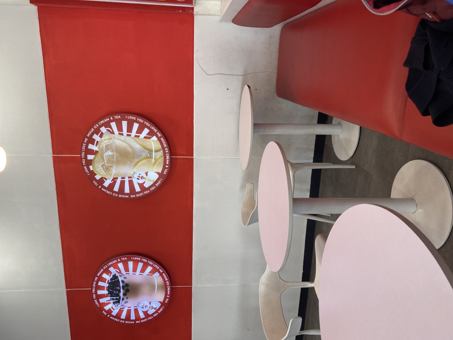 The interior of a restaurant in Kecamatan Piyungan, Indonesia, located at latitude -7.837 and longitude 110.474, features red and white walls with circular advertisements depicting beverages. There are four white chairs and three round, light pink tables. Two chairs are situated at the tables, while one chair is empty. A long red bench with a black sweater lying on top of it is adjacent to a white wall. The wall has a vertical crack in it. The scene is lit by artificial lighting.