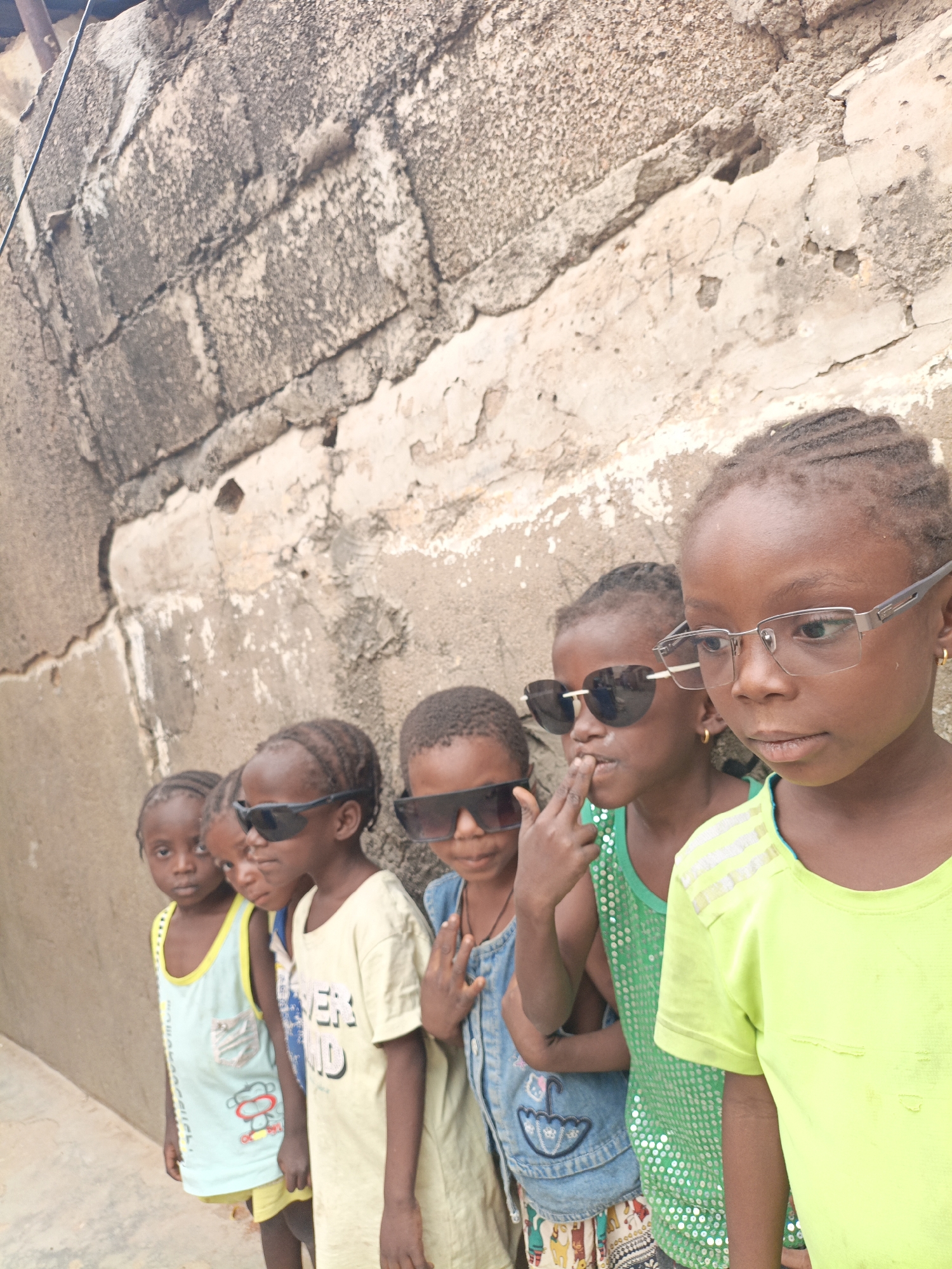 A row of five young Black children stand in front of a weathered, gray brick wall in Gwagwarwa, Nigeria. The children are dressed in casual clothing, with the first three wearing light-colored tank tops and the last two wearing brighter green shirts. Several of the children are wearing sunglasses. The child on the far right wears clear-framed glasses. The wall behind them is textured with aged mortar and exposed brick.
