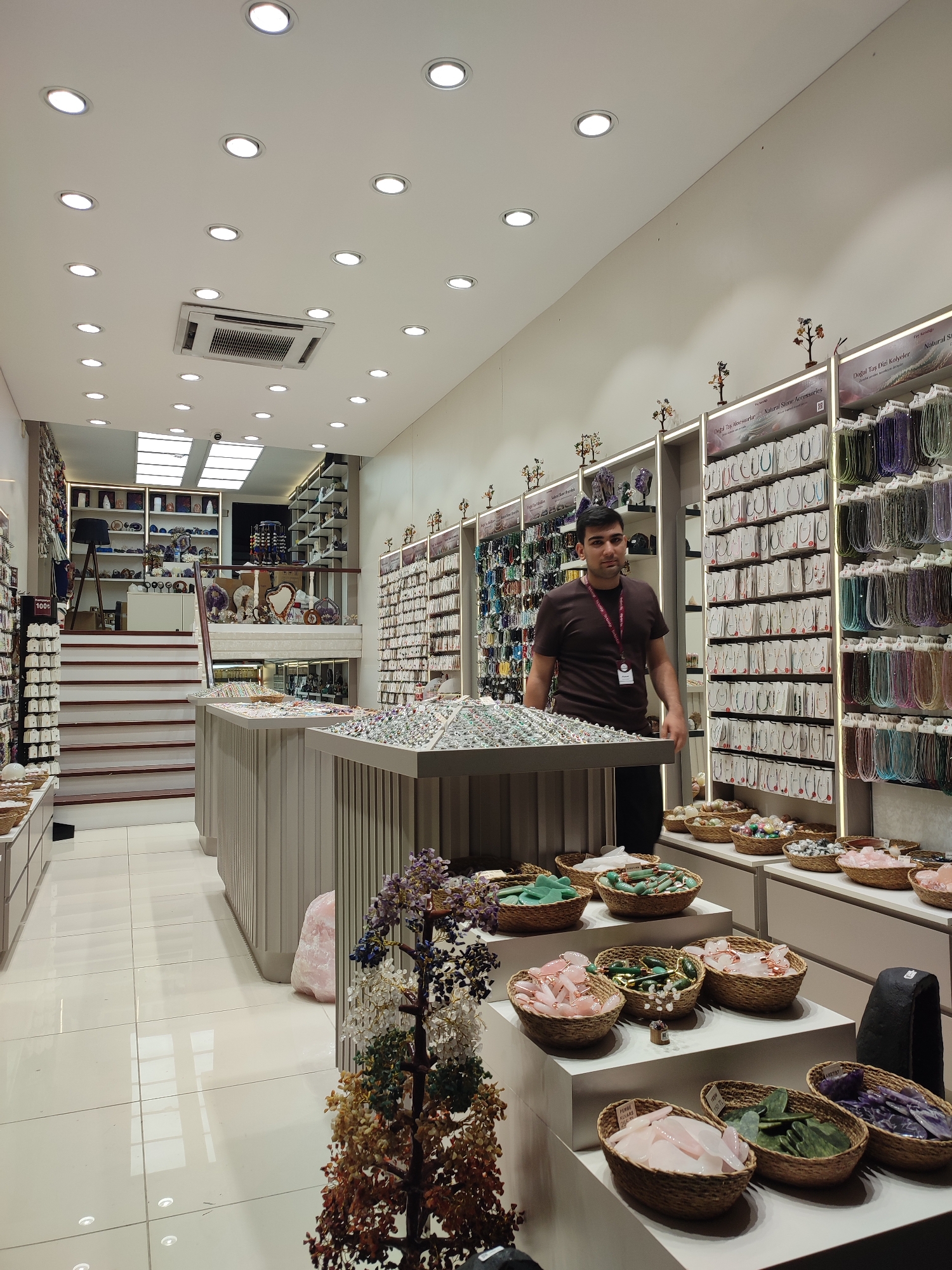 This image depicts the interior of a retail store selling jewelry and accessories, likely located in Kadıköy, Turkey. A man, appearing to be an employee, stands behind a display counter. The store is brightly lit by numerous recessed ceiling lights and features extensive shelving units filled with necklaces, bracelets, and other decorative items. Several display tables are set up in the foreground, showcasing various stone beads and carved stones arranged in baskets. The store has a modern aesthetic with white tiled floors and light-colored display structures. Stairs lead to an upper level where more shelving and display items are visible. The overall impression is of a well-organized and stocked jewelry shop.