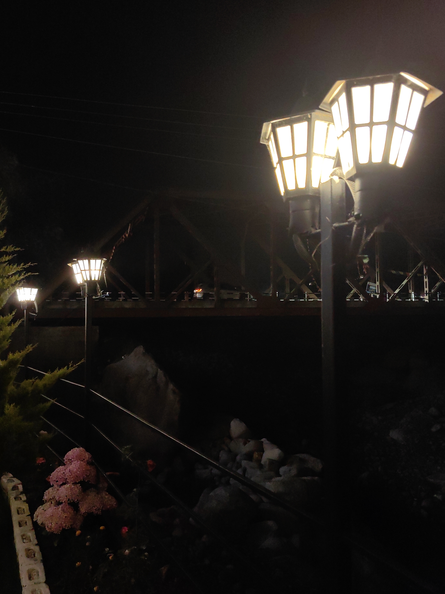 This is a night photograph taken in Kulang, India. The scene is illuminated by a series of ornate, vintage-style street lamps, two of which are prominently featured in the foreground. These lamps have a hexagonal shape with multiple panes, and emit a warm, yellowish light. Behind the lamps, a dark, angular structure, possibly a bridge or a railway trestle, is visible. The bridge's metal framework is silhouetted against the night sky, with hints of distant lights on its surface. In the lower left quadrant of the image, a cluster of pink flowers and some greenery are visible, suggesting a landscaped area. A railing runs diagonally across the bottom of the image, separating the planted area from a rocky, dark embankment. The overall impression is one of a quiet, nocturnal ambiance, possibly in a scenic or historical part of the city.