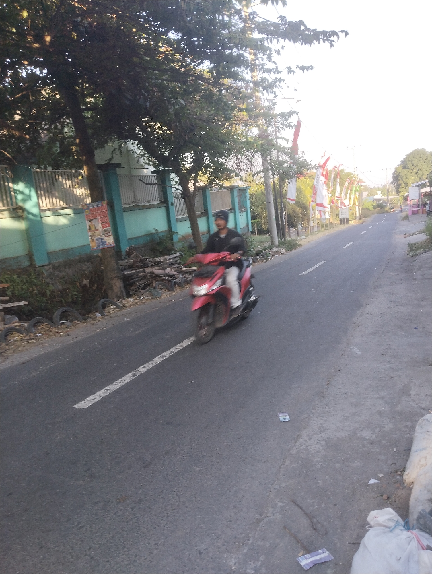 The image depicts a street scene in Wanasaba Daya, Indonesia, likely during daylight hours given the bright sky. A person is riding a red scooter on the right side of a paved road. The scooter is in motion, indicated by slight blurring. The rider is wearing a dark shirt and a helmet. On the left side of the road, there is a turquoise-colored wall with a white fence and some vegetation. Behind the fence, trees are visible. A pole with a poster is also present near the road. Further down the road, there are several white and red flags or banners displayed. The road itself has a white dashed line marking the lane. The ground on the sides of the road appears to have scattered debris, including some tires and trash bags. The overall environment suggests a semi-urban or rural setting.