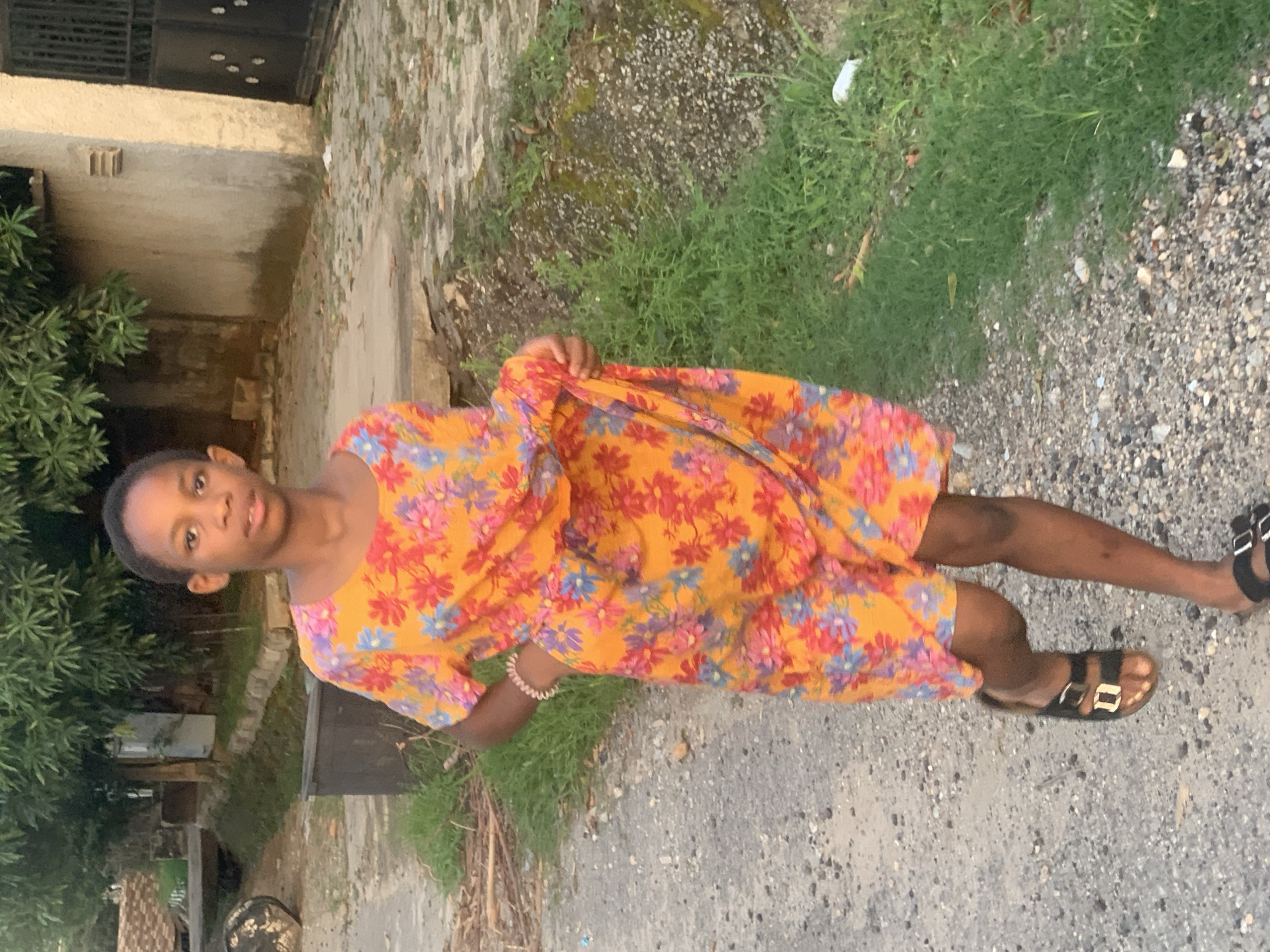 A young person with dark skin stands on a gravel path next to a grassy area. They are wearing a short-sleeved dress with an orange background and a floral pattern of red and blue flowers. They are holding the dress with their left hand, lifting it slightly. The young person is wearing sandals on their feet. Behind them is a building with a dark door and a patch of grass growing against the wall. The scene appears to be in a residential area, possibly in Abuja, Nigeria.