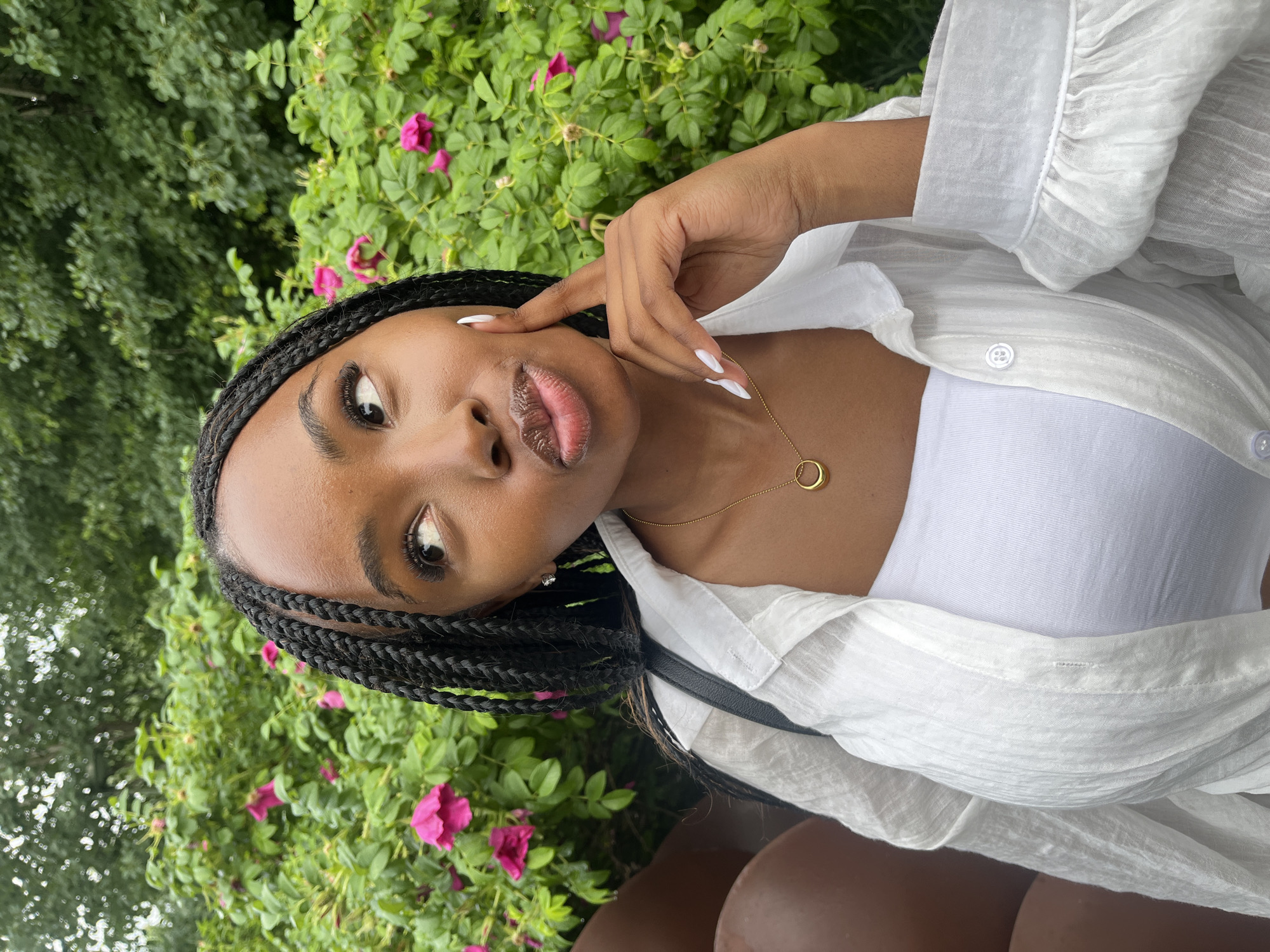 A young Black woman with braided hair is shown from the chest up, posing in front of a bush with pink flowers and green foliage. She is wearing a white button-down shirt open over a white strapless top. A thin gold necklace with a circular pendant is visible. Her right hand is raised, with her index finger touching her cheek. The lighting suggests an outdoor setting. There is no indication of the city of Kazan, Russia.