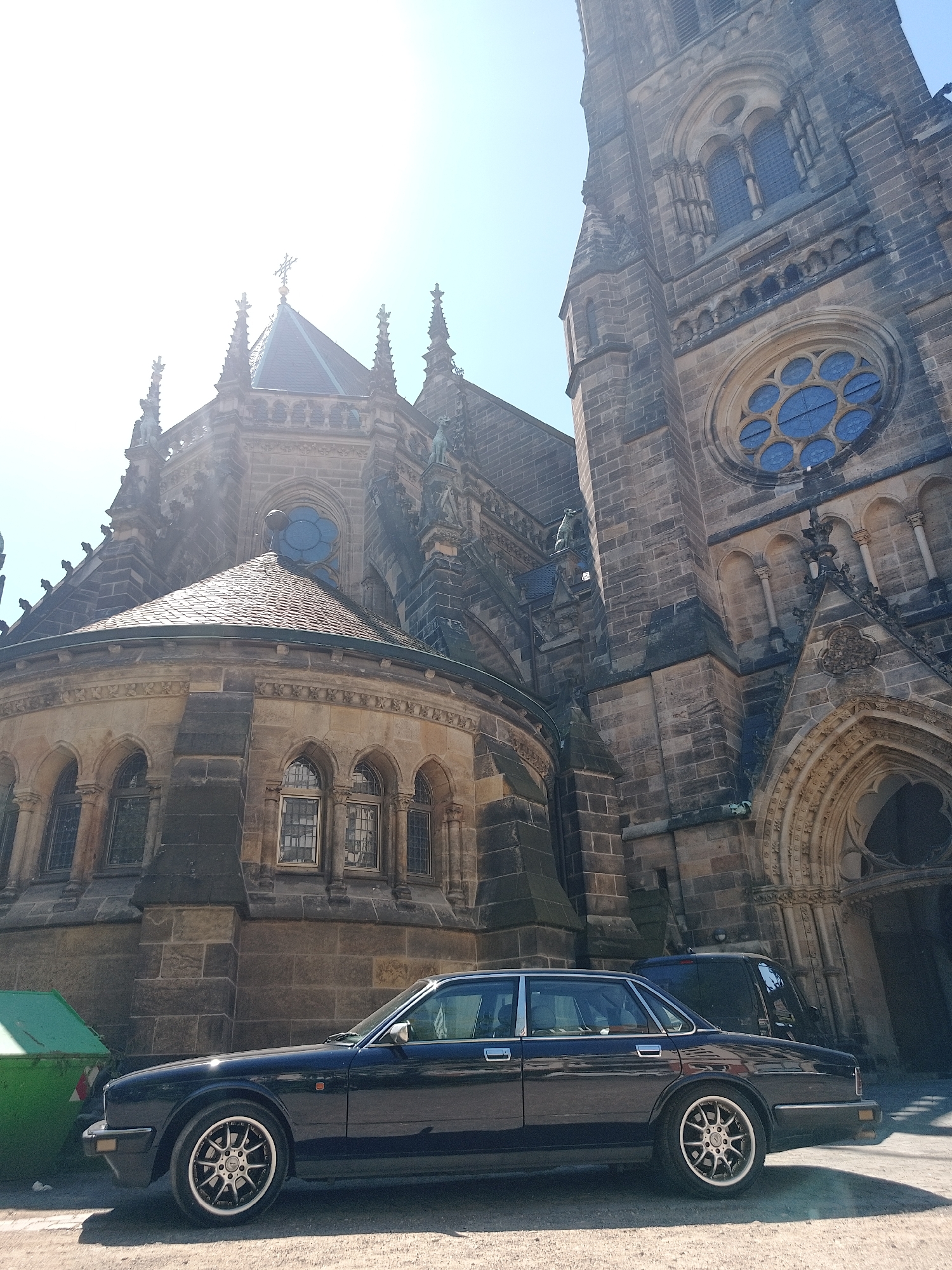 A dark-colored Jaguar sedan is parked on a paved area in front of a large stone church, possibly in Unknown, Unknown. The church features intricate Gothic-style architecture, including a rose window, arched windows, and pointed spires. A green dumpster is visible near the front of the car. Another vehicle is partially visible behind the sedan. The sun is high in the sky, casting shadows and creating a bright, slightly overexposed image.