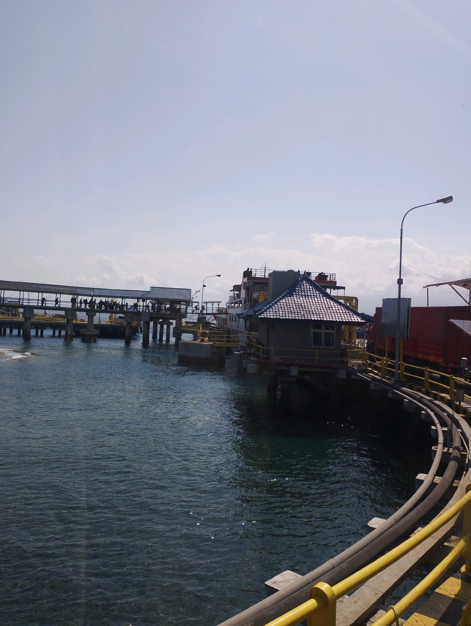 This image depicts a harbor scene in Melaya, Indonesia, on a bright, sunny day. The foreground features a curved yellow railing and large, gray pipes running along a pier. The water, a deep blue with rippling sunlight reflections, occupies the left and lower portions of the image.
Dominating the midground is a long, covered pier with a roof supported by numerous concrete pillars. A group of people, appearing to be passengers or workers, are visible walking along this pier. To the right of the pier, a small, grey building with a dark tiled roof sits on stilts over the water. Beyond this building, parts of a ship are visible, including its deck and superstructure. Further to the right, a red cargo truck or container is parked near the pier, suggesting an active transportation hub. A street lamp on a tall pole stands to the right of the small building. The sky is a pale blue with scattered white clouds. The overall impression is one of a functional, operational port.