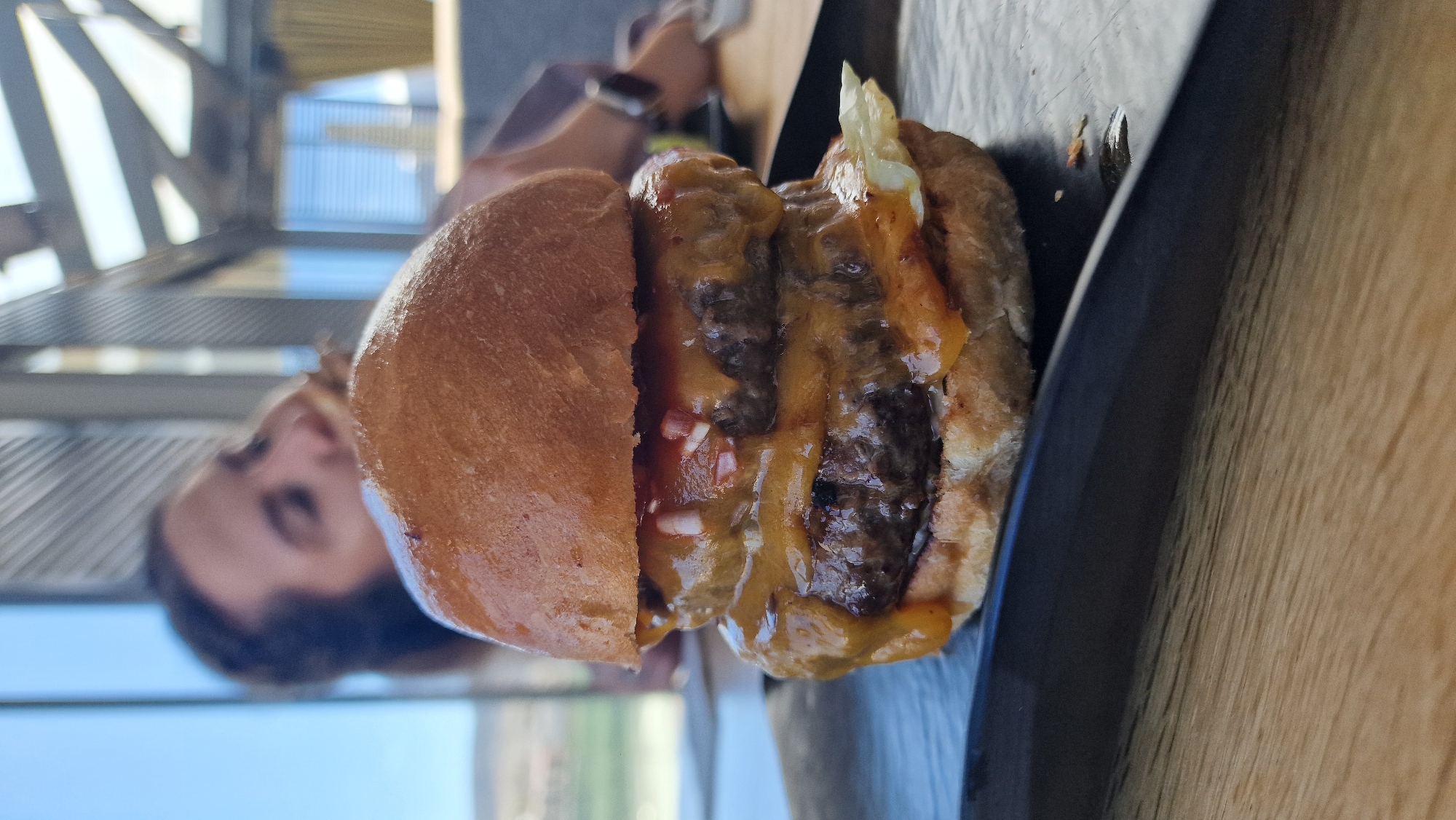 The image depicts a partially consumed double cheeseburger on a dark plate, placed on a light wooden surface. The burger features two beef patties with melted cheese, possibly cheddar, along with visible diced onions and a hint of lettuce. The top bun is halved, revealing the ingredients inside. The burger appears to be outdoors, evidenced by the soft-focus backdrop with a person's face and architectural structures suggesting a building or platform. The overall environment points to an urban setting, possibly a restaurant or patio dining area. The location is in the city of Unknown, Unknown.