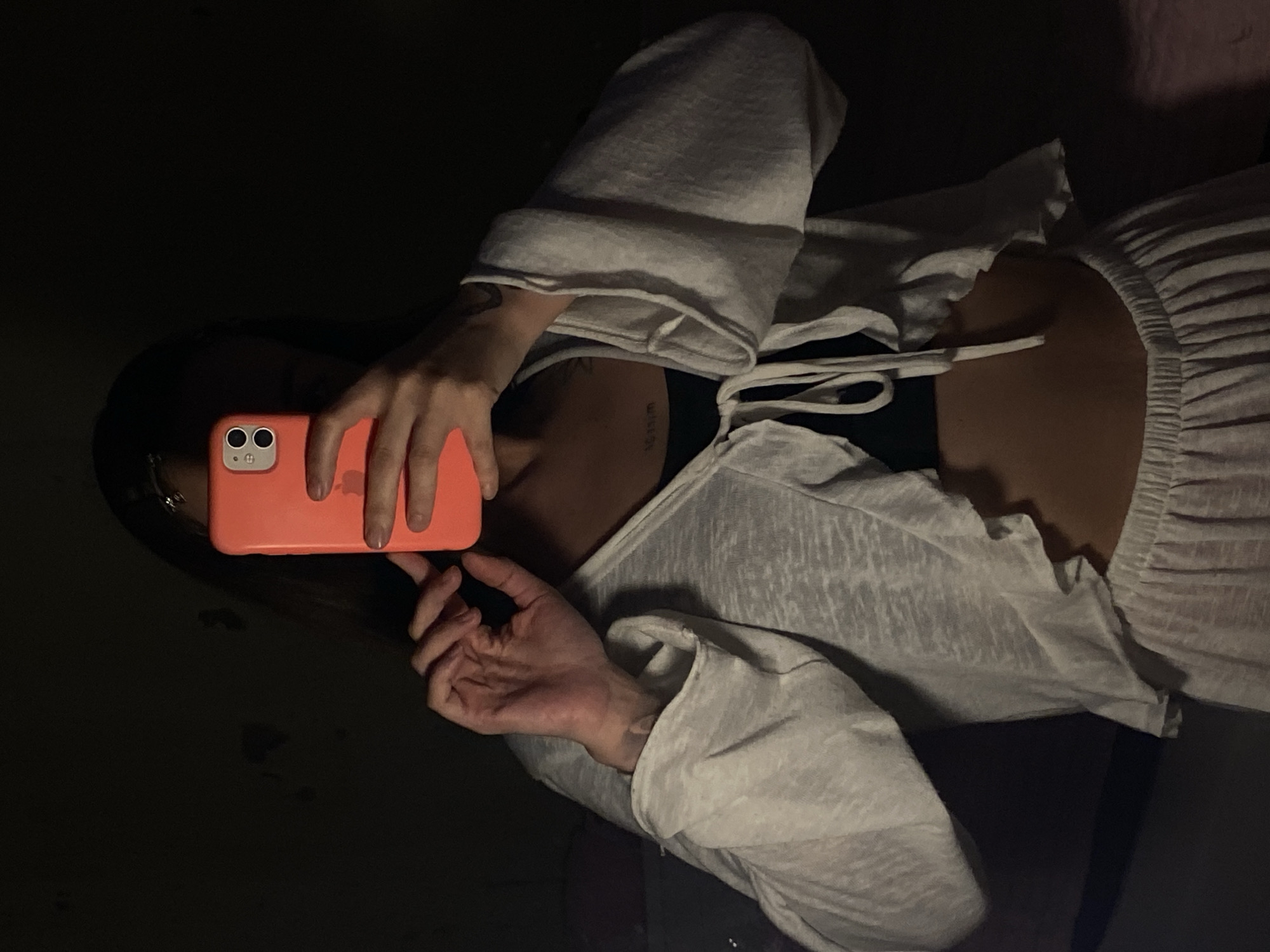 The image captures a partial view of a person, likely female, taking a picture in a dimly lit environment, suggesting nighttime or an indoor setting. The scene is presented from a high angle, looking down towards the person.
The main subject is holding a coral-colored smartphone, likely an iPhone, which has a visible Apple logo and a dual-camera system on its back case. The person's hands are wrapped around the phone, positioned to capture a reflection. They are wearing a light-colored, possibly off-white, long-sleeved top with a textured or ruffled bodice and a tie-front detail visible at the chest. A darker garment, possibly a bra or camisole, is glimpsed underneath the top.
Notable details include an indistinct script tattoo visible on the person's right upper arm (left side of the image). The overall lighting is very low, casting most of the background into deep shadow, with only the immediate vicinity of the person and their clothing being illuminated. No specific location cues are visible within the image itself to confirm it as Votkinsk, Russia, but this is the provided context for the scene. No discernible emotions are visible from the partial view of the person.