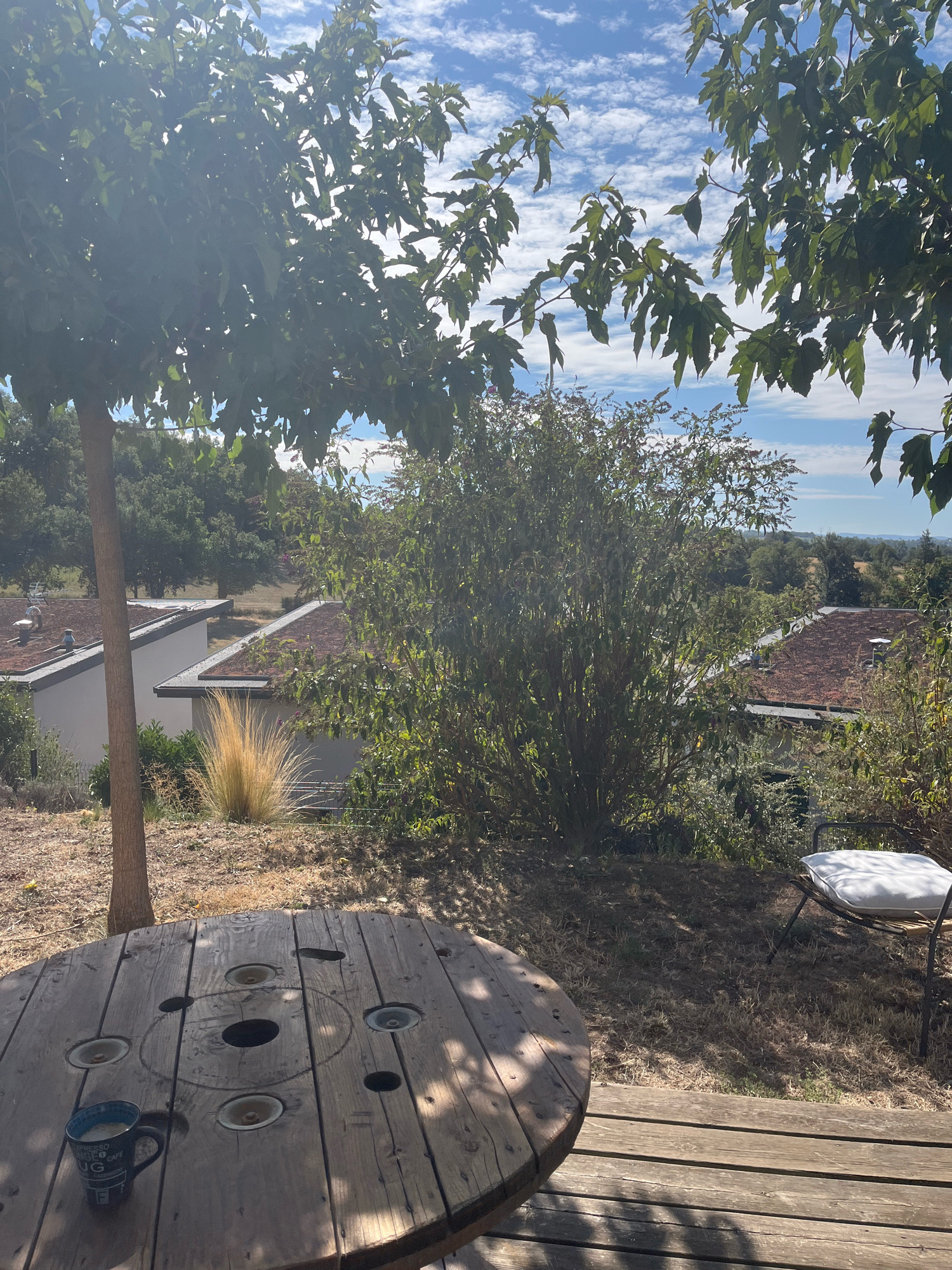 The image captures a tranquil outdoor scene, likely a backyard patio, with a view overlooking a lush, green landscape and a hazy horizon. In the foreground, a rustic wooden table, resembling a repurposed cable spool, dominates the lower portion of the frame. A blue mug, partially filled with a light-colored beverage, sits on the table. To the right of the table, a simple metal chair with a white cushion is positioned, facing away from the viewer. Beyond the patio, the landscape features several buildings with brown, pitched roofs and white walls, some of which appear to have green roofs, suggesting environmentally conscious architecture. Dense foliage, including trees and bushes, frames the scene and extends into the background, interspersed with glimpses of the sky, which is a brilliant blue dotted with fluffy white clouds. The overall impression is one of serene, rural living. The provided location of Chambœuf, France, is consistent with such a picturesque setting.