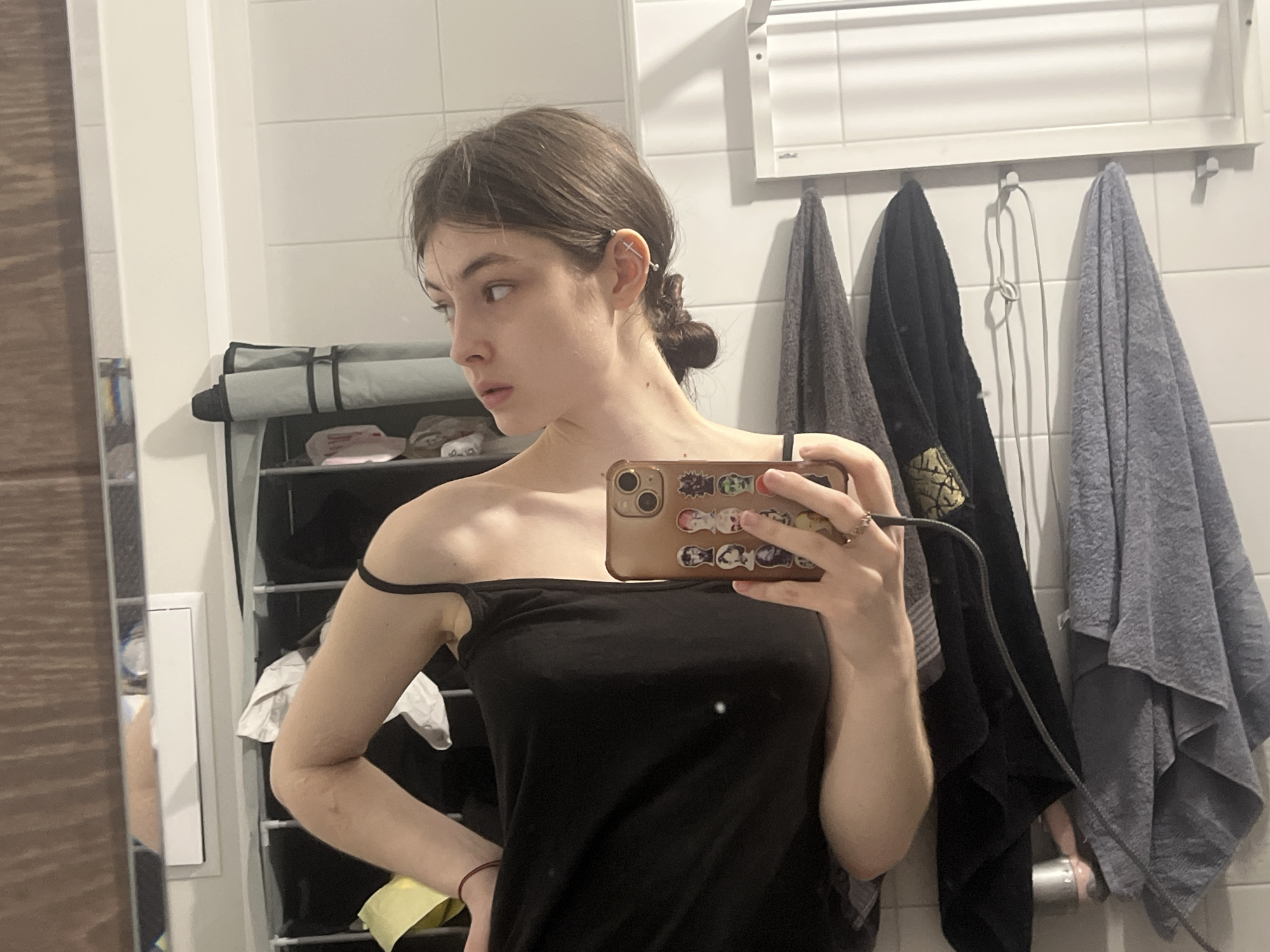 A young woman is taking a selfie in a bathroom with white tiled walls. She is wearing a black tank top with one strap off her shoulder. Her brown hair is pulled back into a bun. She has a piercing in her upper ear. She is holding a phone with a brown case decorated with cartoon characters. Behind her are three towels hanging on a rack: gray, black, and dark gray. A shoe rack is visible on the left, and a wooden door and light switch panel are seen to the left of the shoe rack. The location is not evident from the image.