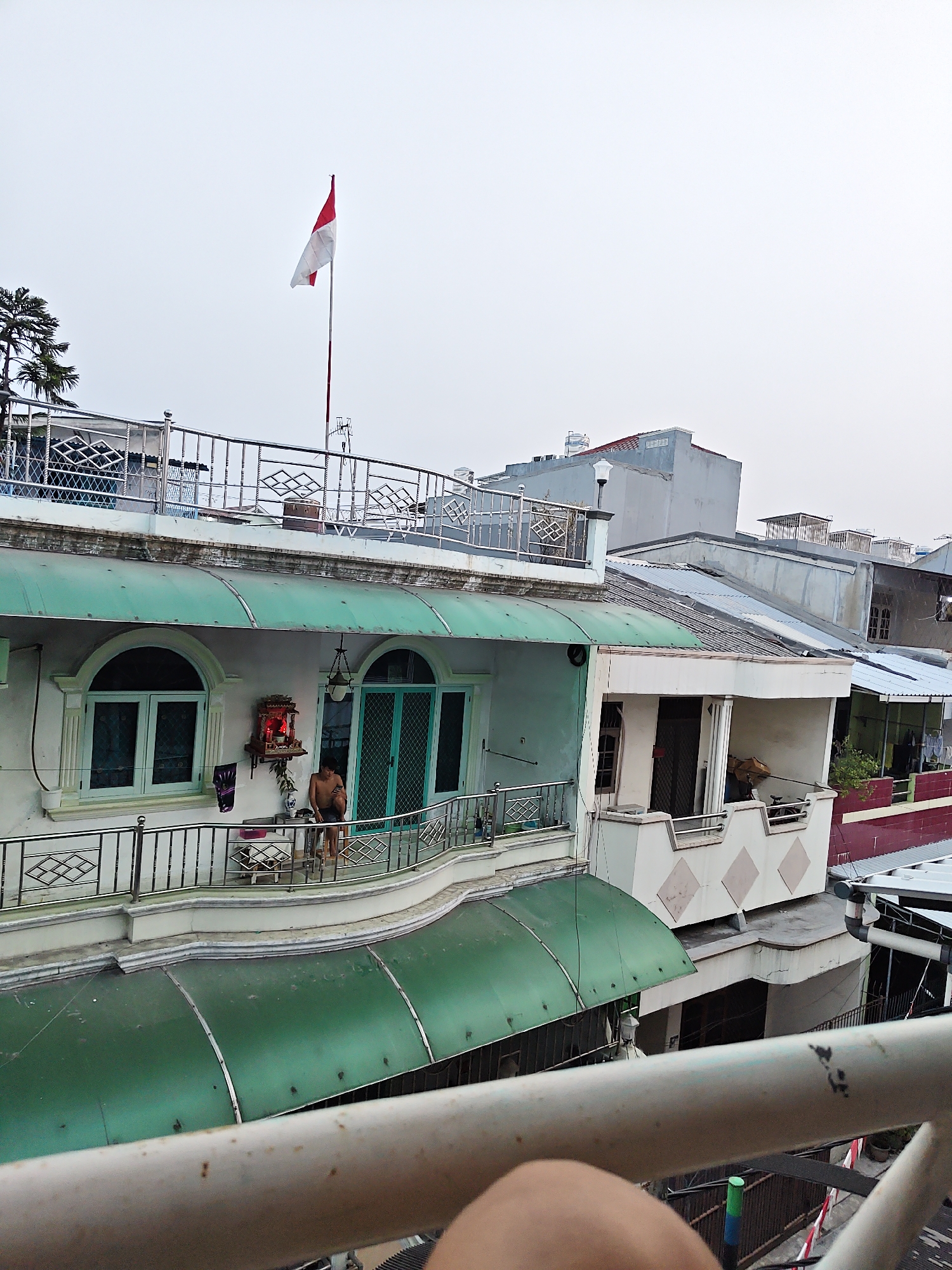 A photograph taken from a slightly elevated perspective captures a multi-story building in North Jakarta, Indonesia. The building features a balcony with a metal railing and a green awning. On the balcony, a person is visible, partially undressed, and appears to be looking at a mobile phone. A small shrine with offerings is also present on the balcony. An Indonesian flag flies from a flagpole atop the building. The background shows other buildings and a cloudy sky, indicative of an urban environment. The overall scene depicts a residential or mixed-use area within the city.