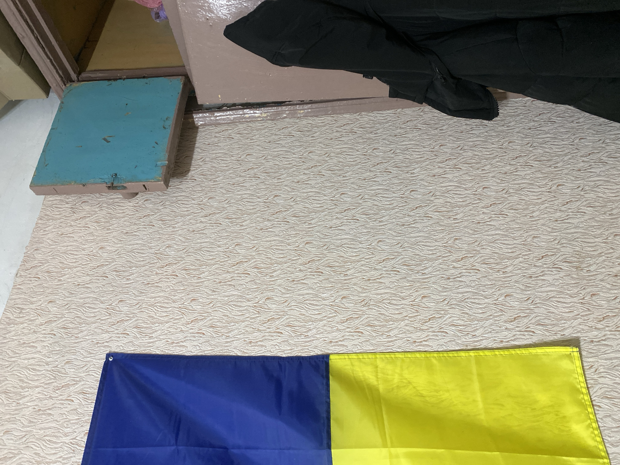 The image shows a room in Poltava, Ukraine, with a blue and yellow Ukrainian flag on the floor. Beside the flag is a closed blue cabinet door. There's also a pile of dark-colored clothes in the background. The floor has a beige textured pattern.
