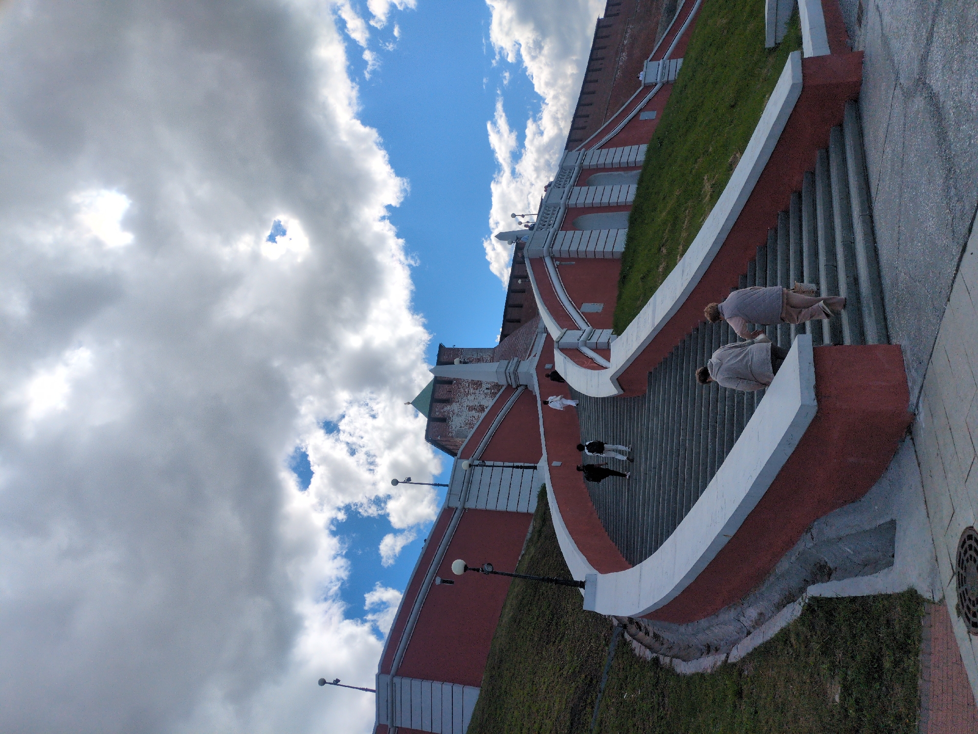 The image depicts a wide, curved staircase constructed from red brick and white concrete, leading up towards a fortified wall. The wall is also red brick and appears to be part of an old structure, possibly a Kremlin, with a green-capped tower visible in the upper left portion of the frame. Lush green grass covers the embankment beside the staircase. Several people are ascending the stairs; two individuals are further up and appear to be walking, while closer to the foreground, two figures are positioned on the stairs, with one leaning forward. The sky above is a vibrant blue, dotted with dramatic white and gray clouds, suggesting a partly cloudy day. Streetlights are positioned along the staircase. The ground adjacent to the stairs is paved with gray material. The scene captures an outdoor setting in Nizhny Novgorod, Russia, during daylight hours with favorable weather conditions.
