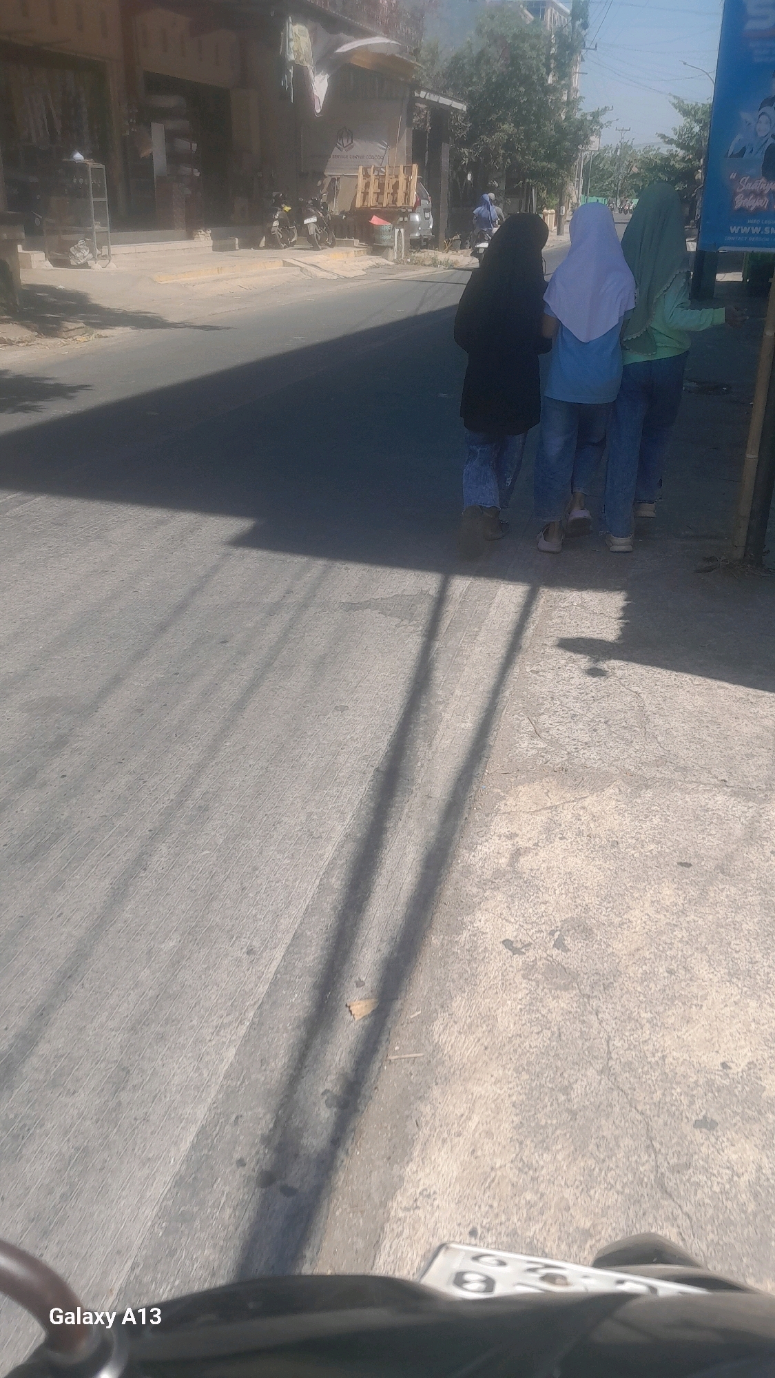 Three individuals, two women and one person whose gender is not discernible, are walking away from the camera on a paved road. The person on the left wears a black hijab and blue jeans. The person in the middle wears a white hijab and blue jeans. The person on the right wears a green hijab and blue jeans. They are walking past a building on the left side of the frame which appears to be a shop or business. In the background, there are trees, a clear sky, and a billboard on the right. The scene is lit by bright sunlight, casting distinct shadows on the road. The lower portion of the image shows a portion of a motorcycle's dashboard with a license plate. This image was taken in Kirig, Indonesia.