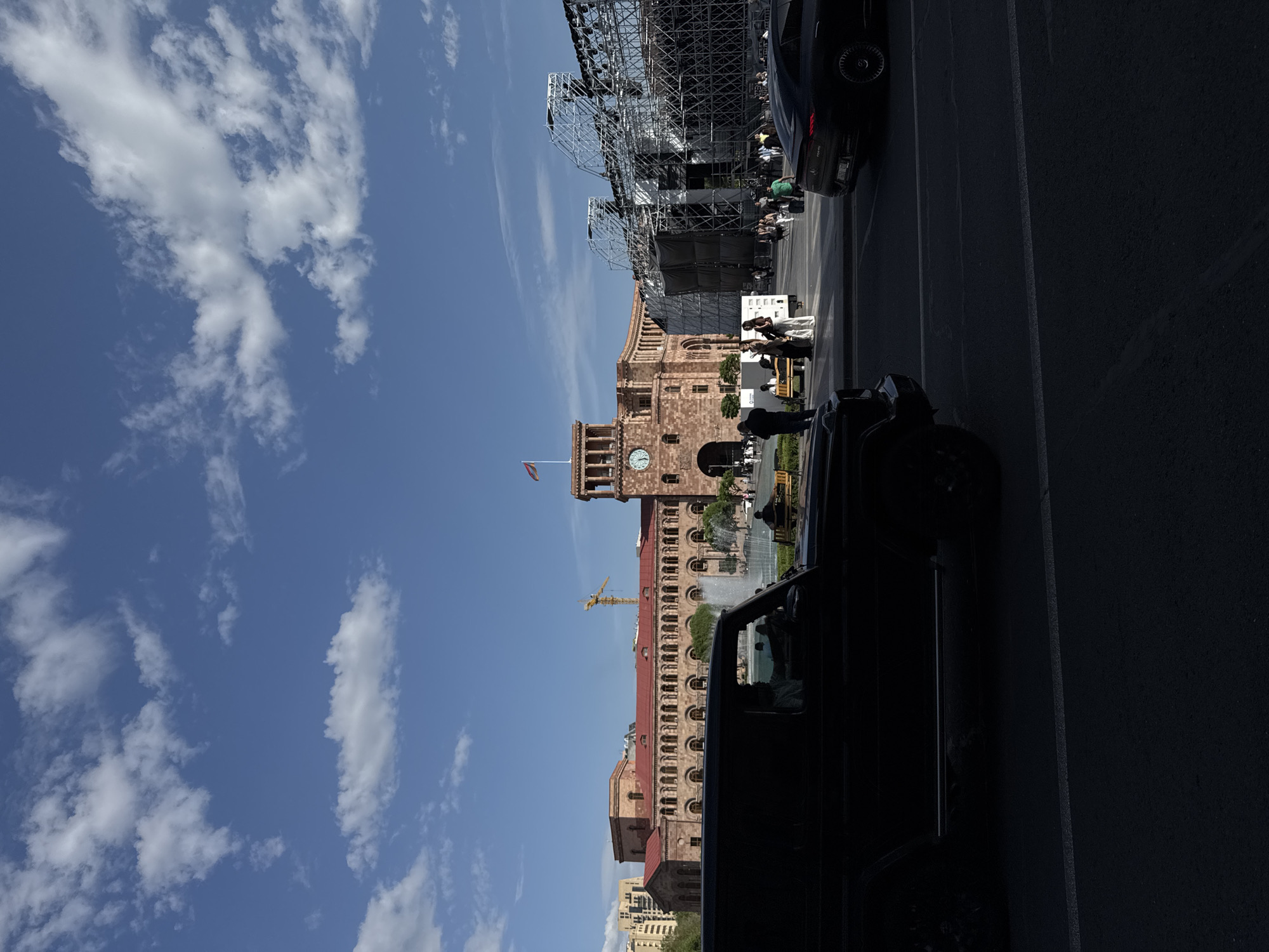 The image shows a daytime street scene in Yerevan, Armenia. The sky is blue with scattered white clouds. A large stone building with a clock tower and an Armenian flag on top dominates the central background. There are several people, mostly indistinct, near the building. A large metal stage structure is visible to the right of the building. Several cars are parked and driving in the foreground, partially obscuring the view. The road surface is asphalt with white lane markings. The overall light suggests it's a sunny day.