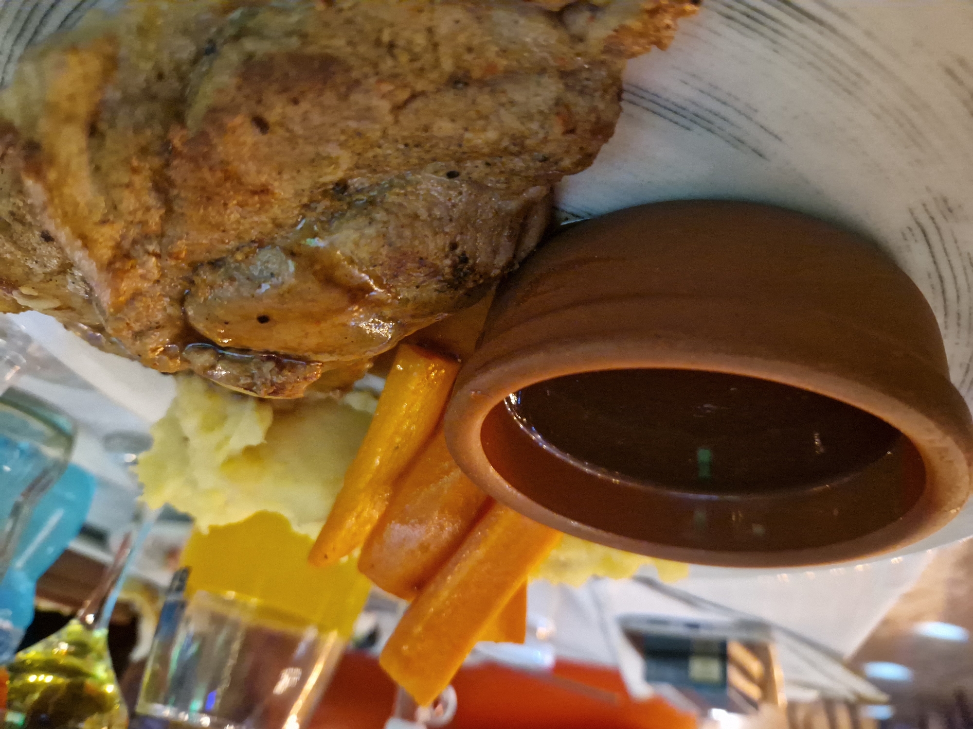 A close-up shot captures a plate of food, likely a meal in a restaurant. The dish features a piece of steak, generously coated in a dark, glossy sauce. To the left, a small, rustic brown bowl holds more of the same sauce, its surface reflecting the overhead lights. Beside the steak and sauce bowl are mounds of mashed potatoes and several pieces of cooked carrots, cut into rectangular shapes. The food is served on a white plate with a subtle, grey, brushed pattern. In the background, out of focus, a glass with a yellow liquid, possibly juice or wine, stands on a table, suggesting a dining setting. The overall impression is a hearty, prepared meal ready to be consumed, possibly in a restaurant in Tankovo, Bulgaria, given the context.