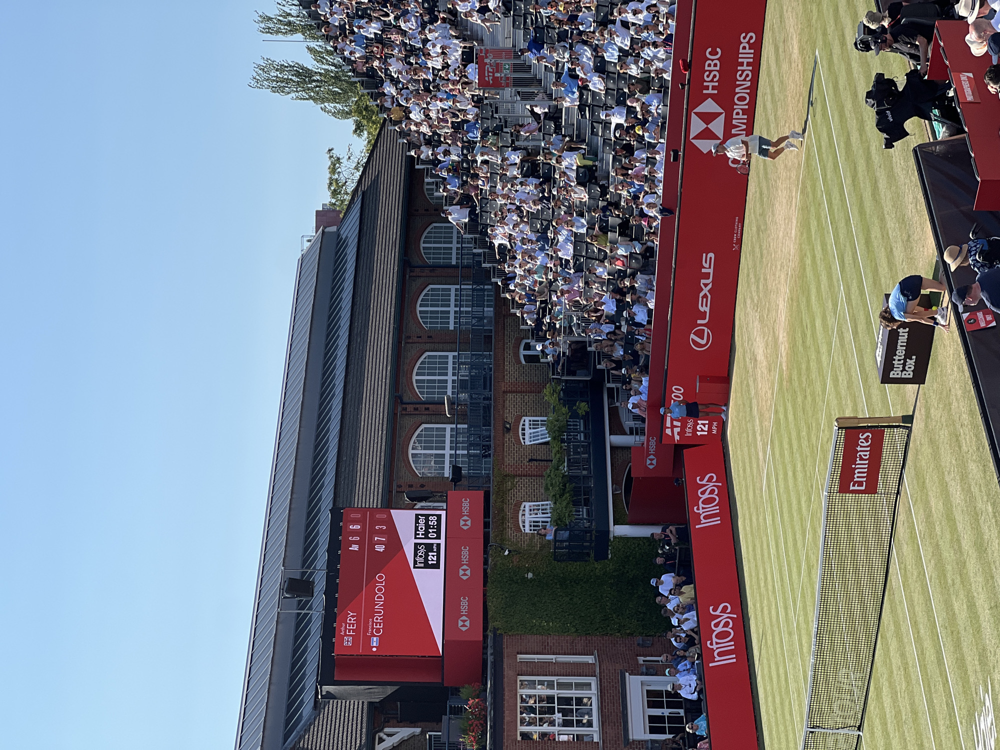 This is a medium-shot, eye-level, outdoor, daytime photograph of a tennis match on a grass court. The scoreboard is prominently displayed on a large red screen mounted above a building with a dark grey roof and red brick facade. The scoreboard shows "Arthur FERY" and "Francisco CERUNDOLO" as players, with a score of "Av 6 6 0" for Fery and "40 7 3 0" for Cerundolo. Below the player names are sponsor logos for "Infosys" and "Haier," displaying "121 MPH" and "01:58."
In the foreground, the net of the tennis court is visible. To the left, a red banner with "Infosys" logos runs along the edge of the court. To the right, a player in a white shirt and blue shorts is about to serve, holding a tennis racket. Spectators are seated in tiered bleachers on the right side of the image, most wearing light-colored clothing. Another player, wearing a blue shirt and dark shorts, is on the left side of the court, bent over to retrieve a tennis ball. A camera on a tripod is positioned near the court, and other personnel are visible in the background. The sky is clear and blue.