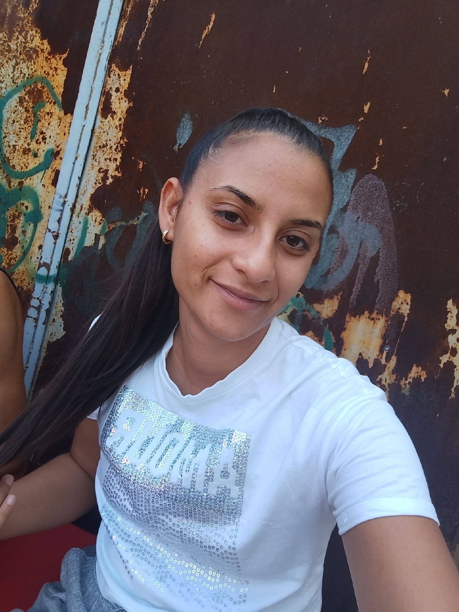 A young woman with dark hair pulled back in a ponytail, wearing a white t-shirt with a sequined design and a gold hoop earring, smiles at the camera. She is positioned in front of a rusted metal surface with faded graffiti. The image appears to be a selfie taken outdoors. There is no information available regarding the location in the city of Yambol, Bulgaria.