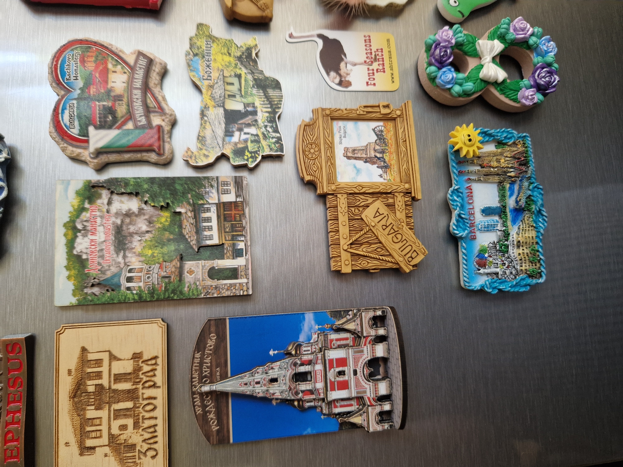 The image displays a collection of ten distinct refrigerator magnets adhered to a brushed metallic surface.
Positioned in the upper left, a dark brown rectangular magnet bears the raised red text "EPHESUS." To its right, a light-colored wooden rectangular magnet is engraved with a two-story traditional house and the Cyrillic text "ЗЛАТОГРАД" (Zlatograd) below the illustration. Above the center-right, a die-cut magnet depicts a landscape with a monastery tower and rock formations, inscribed with "ДРЯНОВСКИ МАНАСТИР" (Dryanovski Manastir) and "Dryanovo Monastery." Adjacent to this, a heart-shaped magnet features a stylized Bulgarian flag, an image of houses and trees, and the text "BULGARIA," "Bachkovo Monastery," and "БАЧКОВСКИ МАНАСТИР" (Bachkovski Manastir).
In the middle-left area, a tall, arched rectangular magnet presents a 3D relief of the multi-domed Shipka Memorial Church, rendered in red, white, and gold, against a blue sky background. Text above the church reads "ХРАМ-ПАМЕТНИК" (Church-Monument), "РОЖДЕСТВО ХРИСТОВО" (Nativity of Christ), and "ШИПКА" (Shipka). To its right, a map-shaped magnet outlines Bulgaria, displaying a traditional white house within a green landscape, with the text "Боженци" (Bozhentsi). Below the Shipka Memorial Church magnet, a rectangular wooden magnet, designed as an opening gate, frames an image of the Shipka Peak Monument (a stone tower with cannons), inscribed "Shipka Peak Bulgaria." A wooden panel on its left is carved with "BULGARIA." Next to the Shipka Peak magnet, a die-cut magnet in the shape of an ostrich, colored white, black, and brown, carries the text "Four Seasons Ranch" and "www.shtraus.com."
At the bottom left, a rectangular magnet with a blue braided border depicts a Barcelona cityscape, including the Sagrada Familia, modern skyscrapers (such as the Agbar Tower), a body of water, and a smiling yellow sun in the upper right. This magnet is labeled "BARCELONA." To its right, a cream-colored magnet is shaped as the number 8, decorated with a wreath of blue and purple roses and a white bow centered within the upper loop of the '8'. A small green object, possibly another magnet, is partially visible on the far right edge.